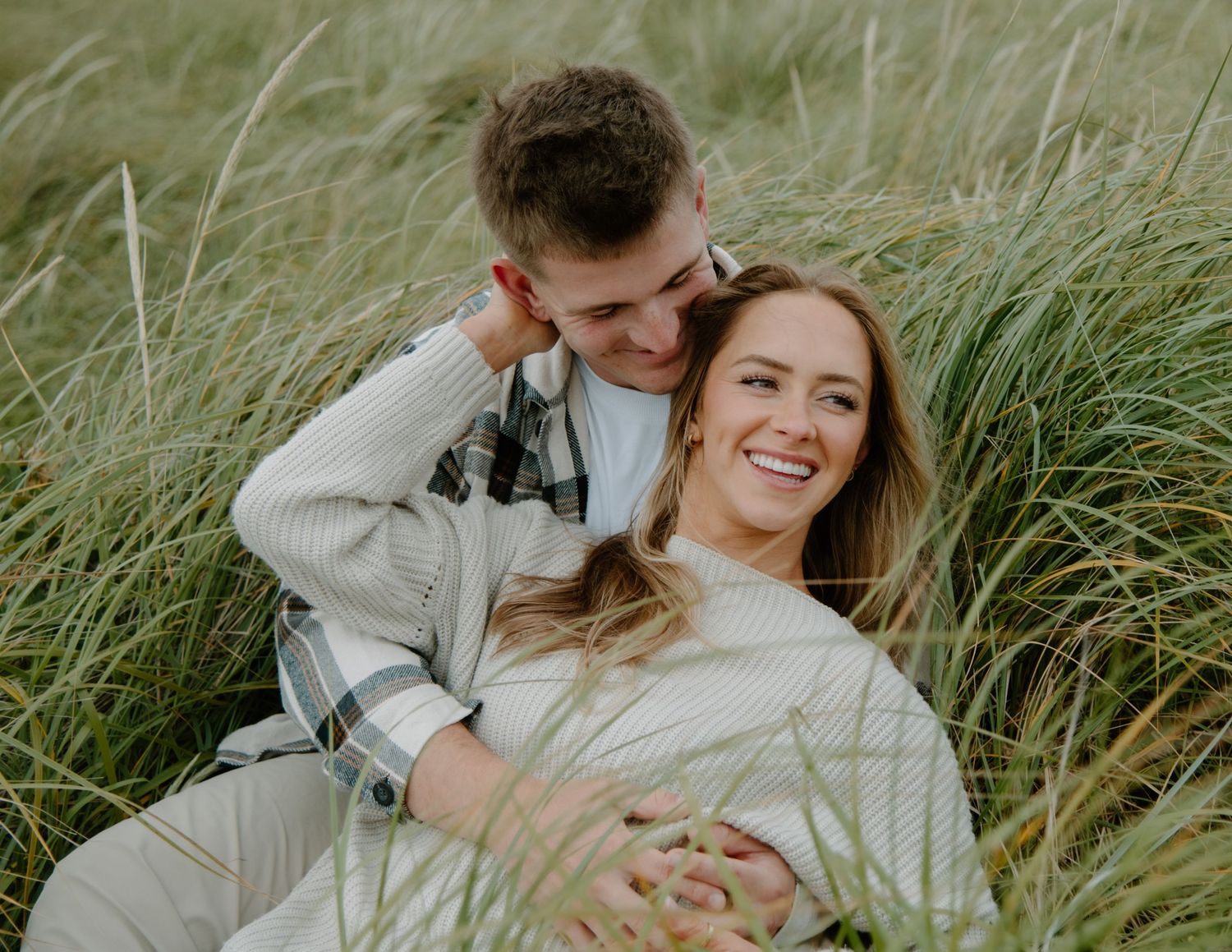 Romantic couple embraces while sitting in tall grass on a sunny day on Oregon Coast. Calgary Lifestyle and Engagement Photographer.