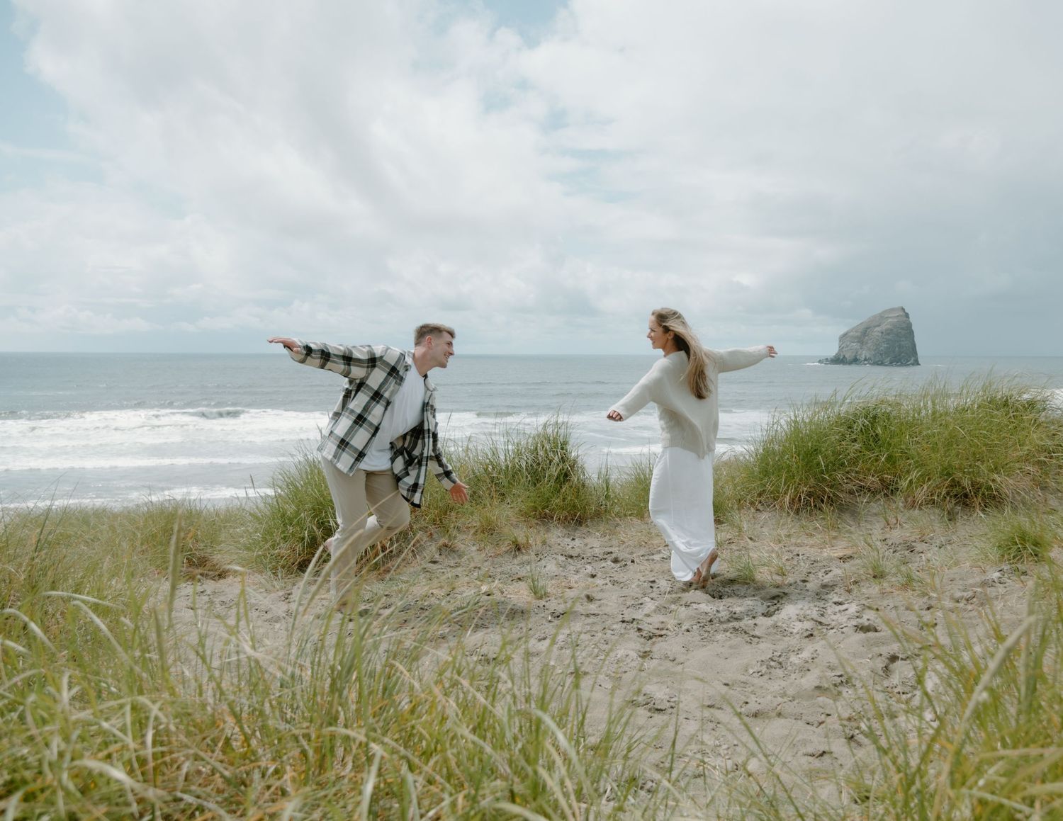 Couple playing and running through coastal dunes with Haystack Rock visible in the distance at Pacific Beach on Oregon Coast.