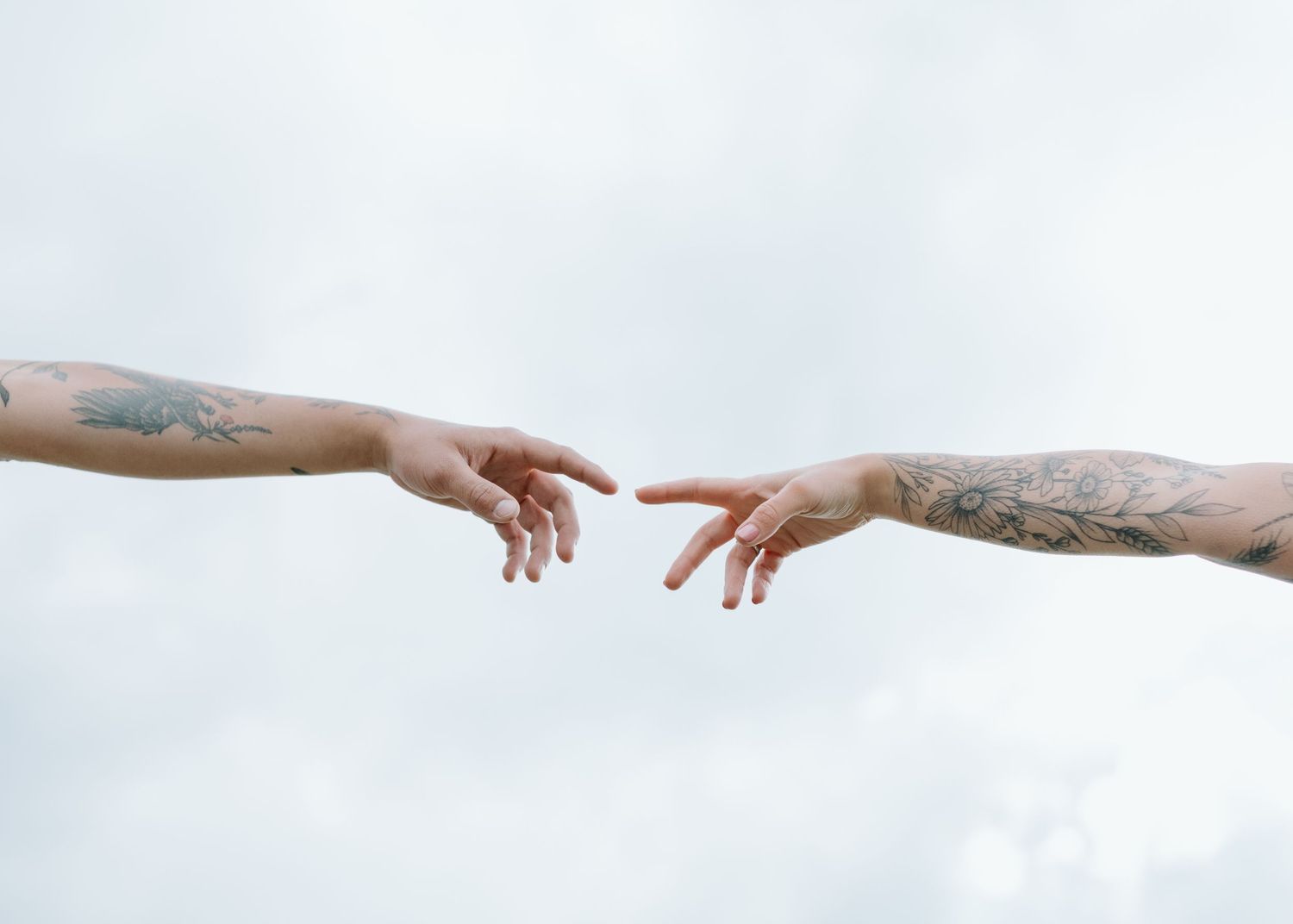 Two tattooed arms reach toward each other against white sky in recreation of Michelangelo's Creation. Calgary Alberta Engagement Photographe