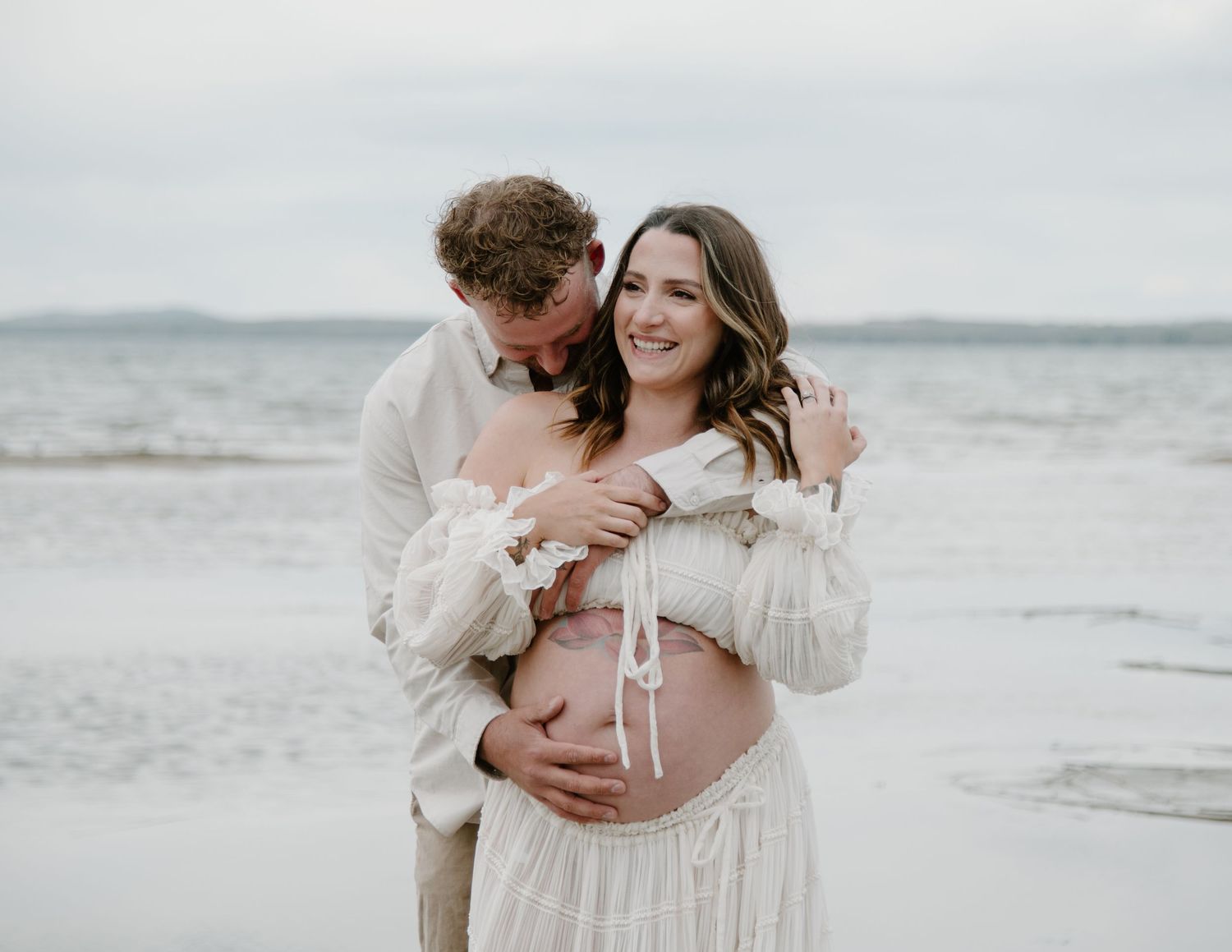 A tender maternity moment captured on a cloudy beach with a couple in light coloured outfits. Calgary Alberta Maternity Photographer.