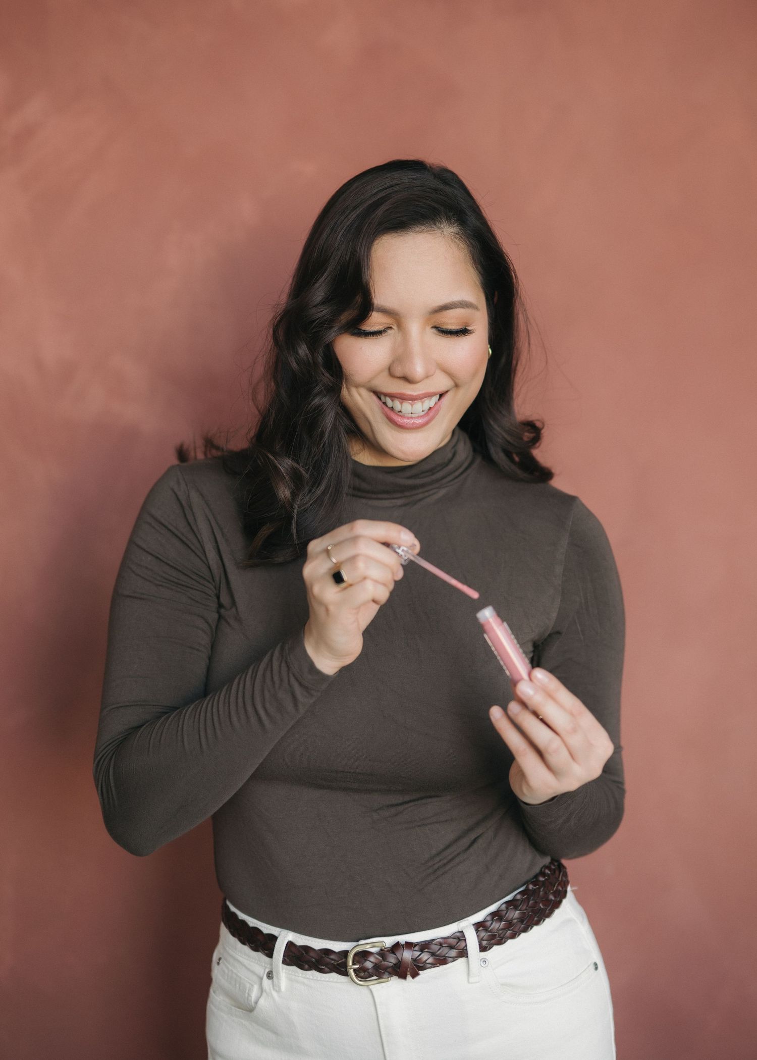 A sequence of photos shows someone in a gray sweater applying pink lip gloss and smiling against a reddish-brown background.