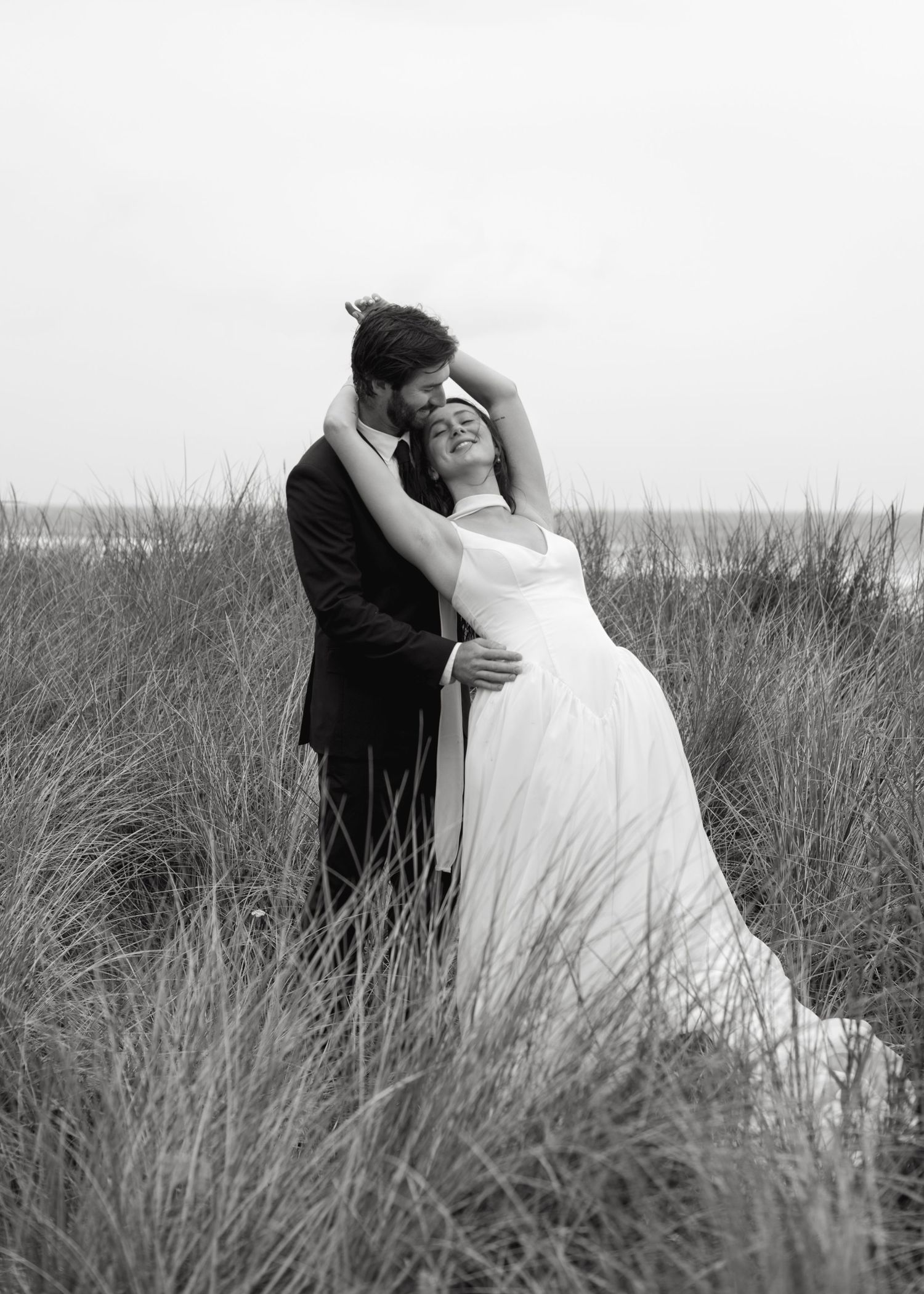 A romantic black and white wedding portrait of a couple embracing in tall grass with the bride wearing a flowing white dress.