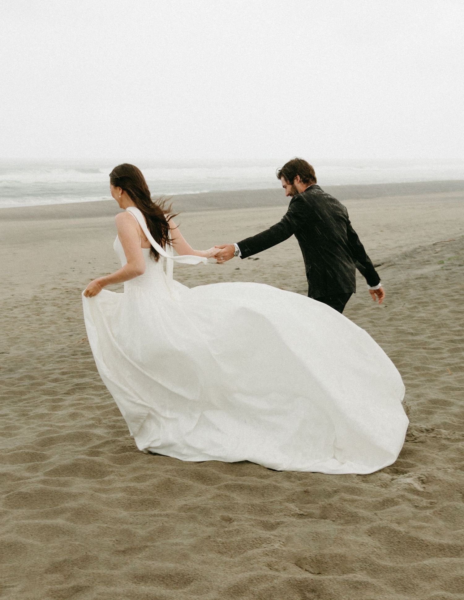 A couple in wedding attire dance on a misty beach as the bride's white dress billows dramatically in the wind.