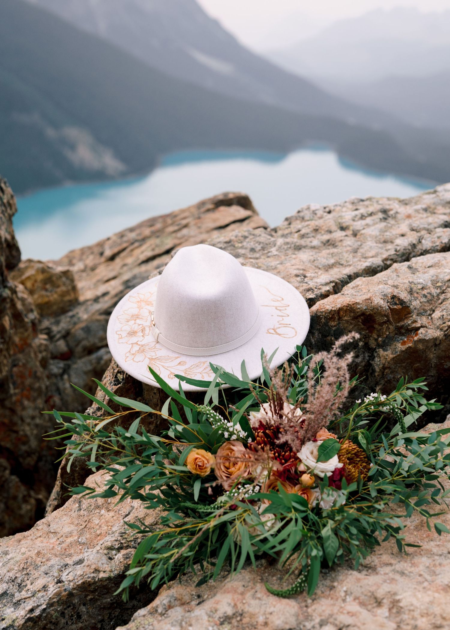 A white wide-brimmed hat and floral bouquet arranged on rocky cliffs overlooking turquoise lake waters in misty mountains.