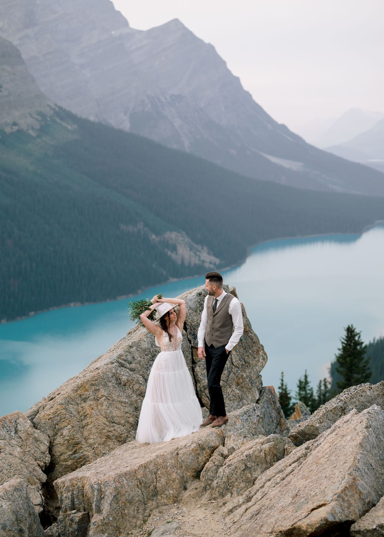 Couple in wedding attire share intimate moment on mountain ledge with stunning turquoise lake and mountains in background.