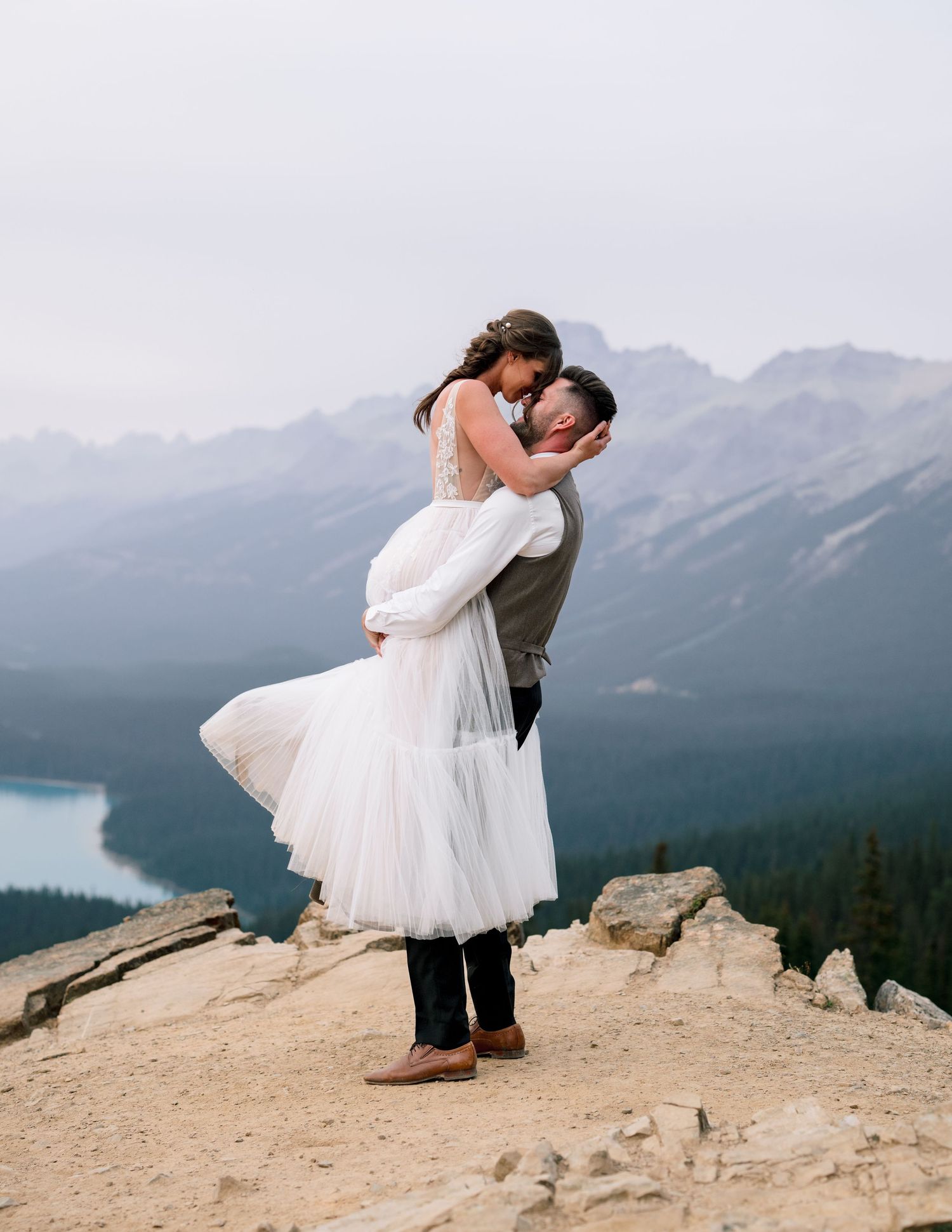 Newlyweds embrace on a mountain clifftop overlooking scenic mountain vistas and a lake below.