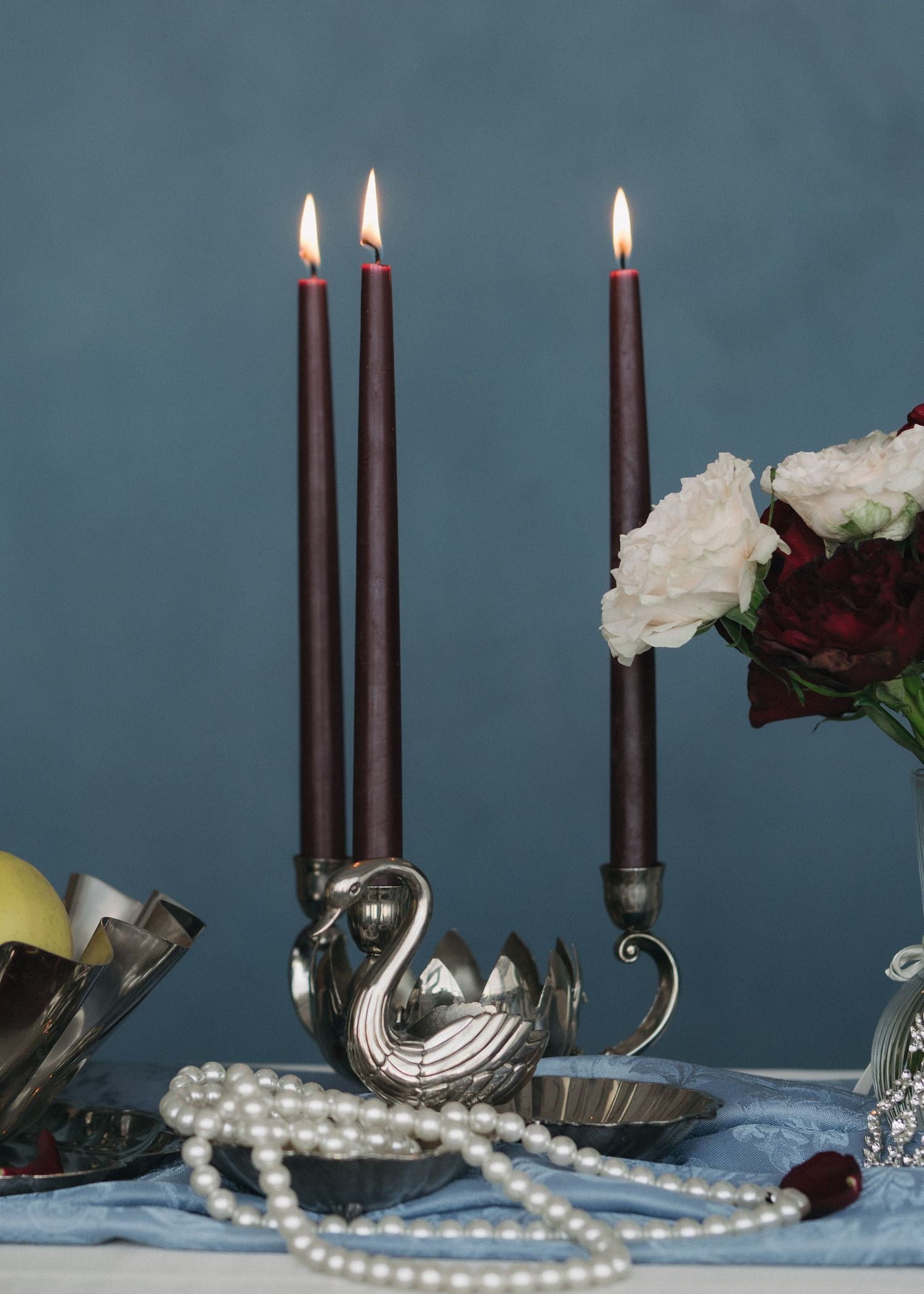 Elegant table setting with lit black candles, silver teapot, and pearl beads against a moody blue-gray background.