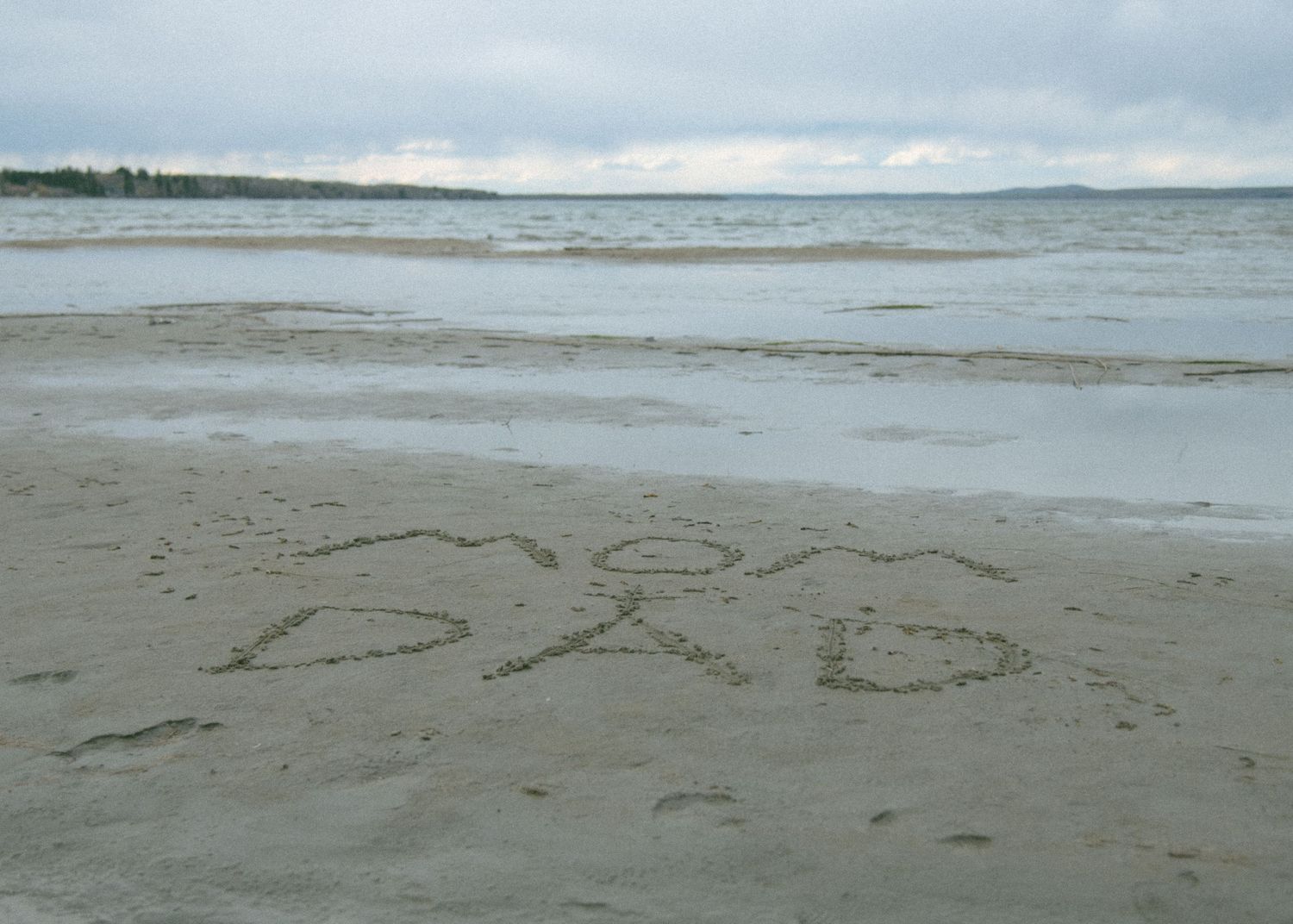 Text written in sand on a beach with cloudy skies and ocean waves. Calgary Alberta Maternity Photographer.