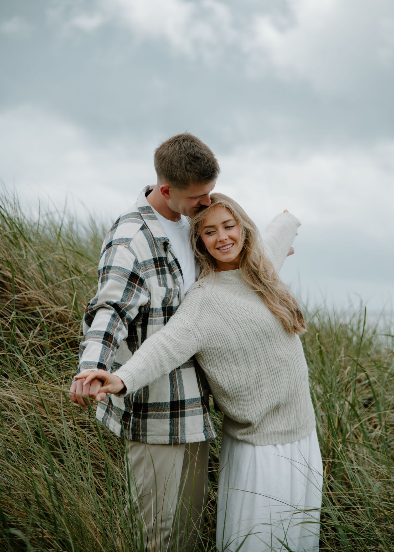 Couple shares intimate moment among tall swaying grass on cloudy day on Oregon Coast. Calgary Lifestyle and Engagement Photographer.