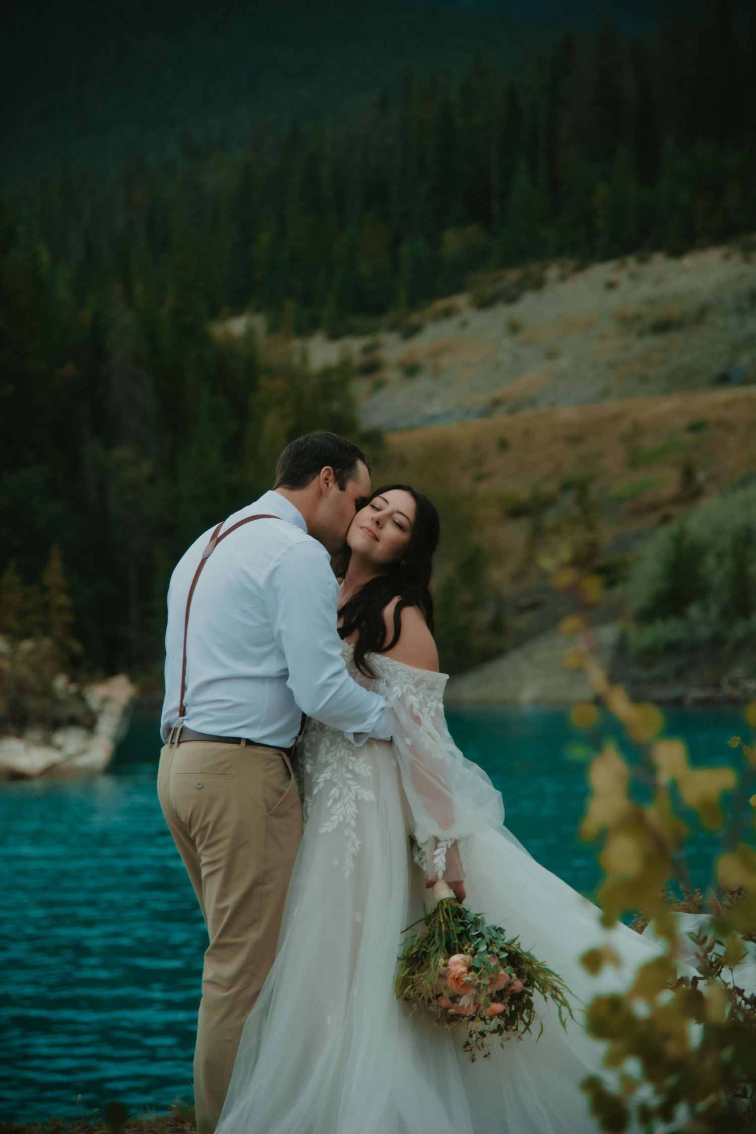 Newlyweds share an intimate moment by a turquoise mountain lake with evergreen forest in background. Calgary Wedding Photographer