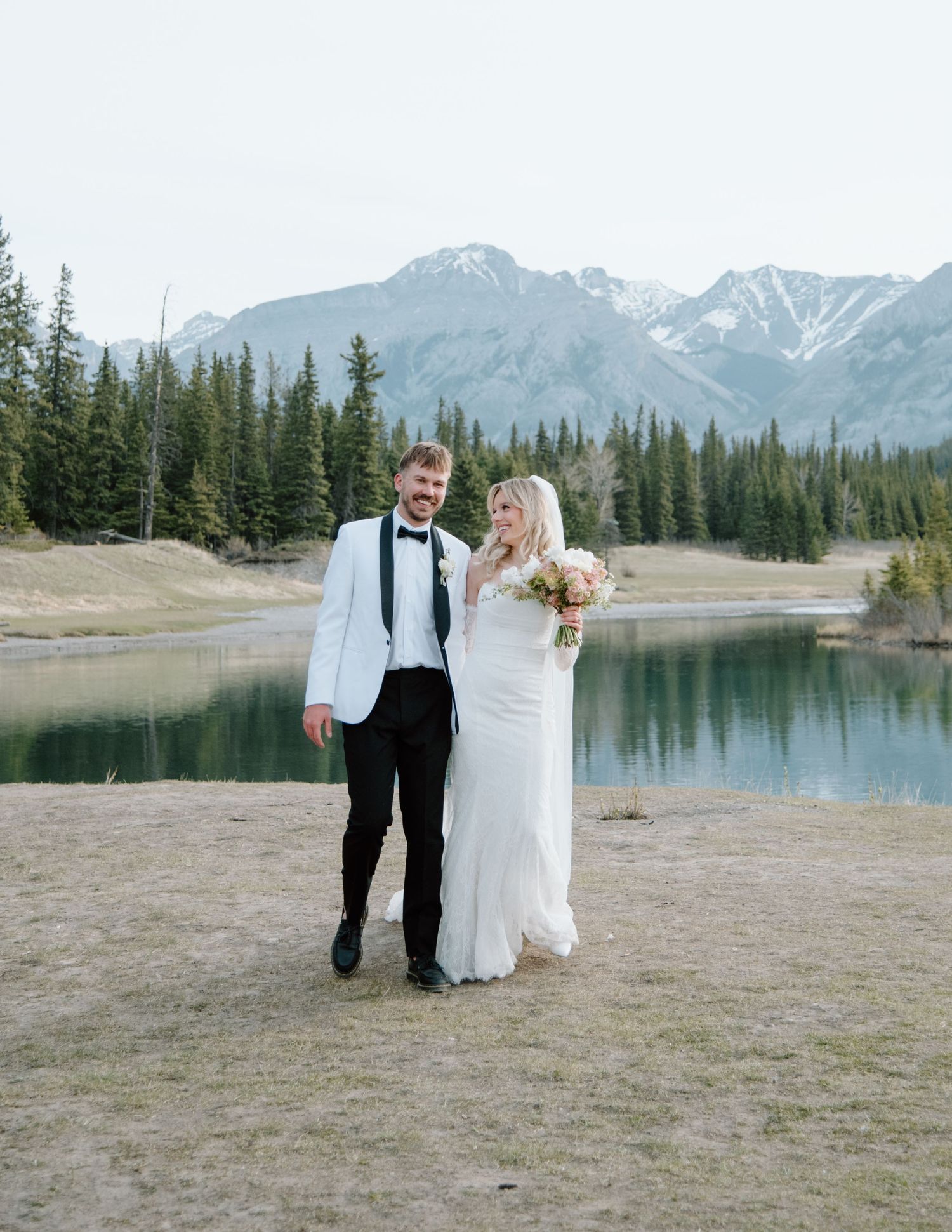 Newlyweds pose together by scenic mountain lake with snowy peaks in background. Calgary Wedding Photographer
