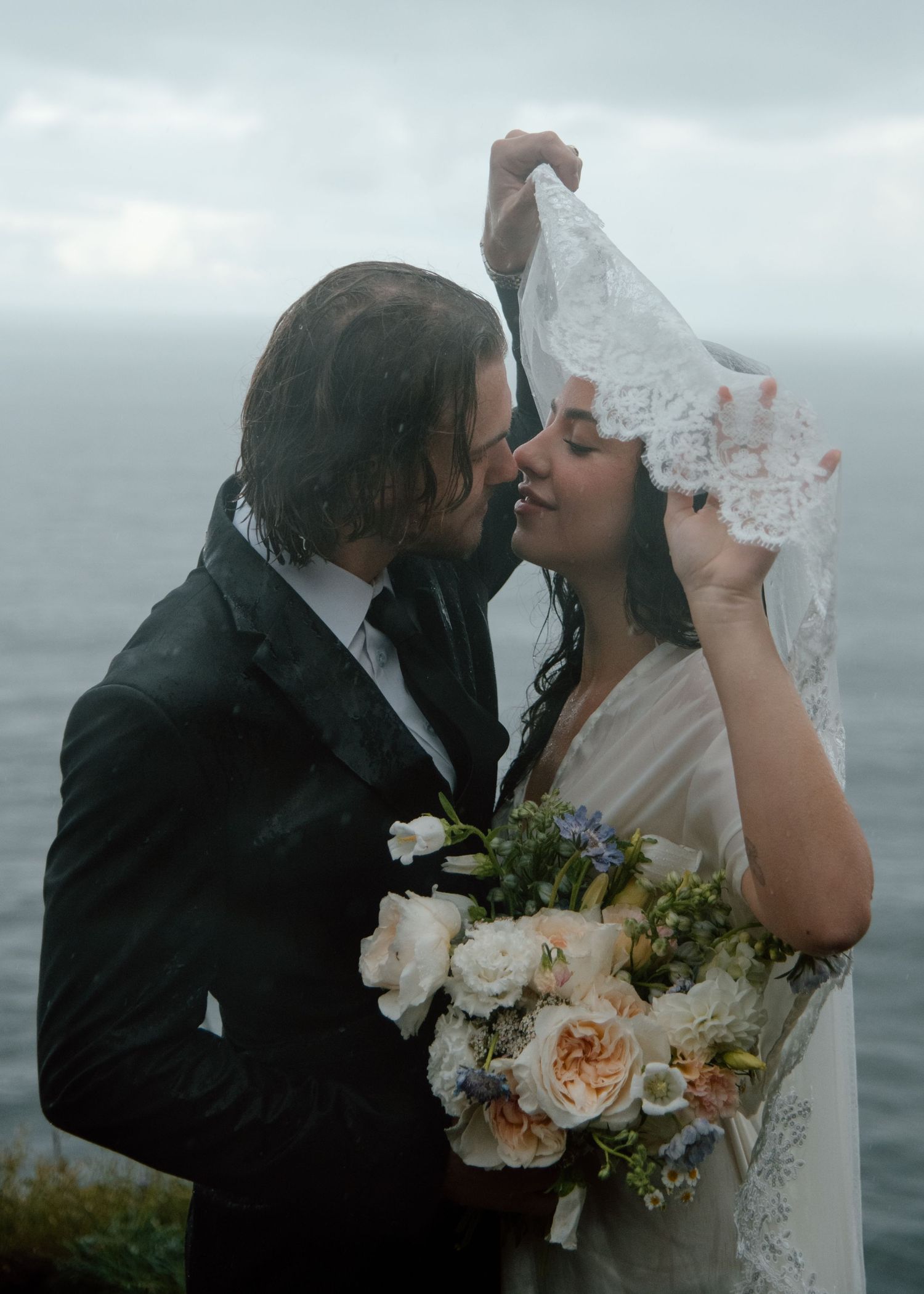 Romantic wedding couple embraces during overcast coastal photo session with peach and white floral bouquet. Calgary Wedding Photographer