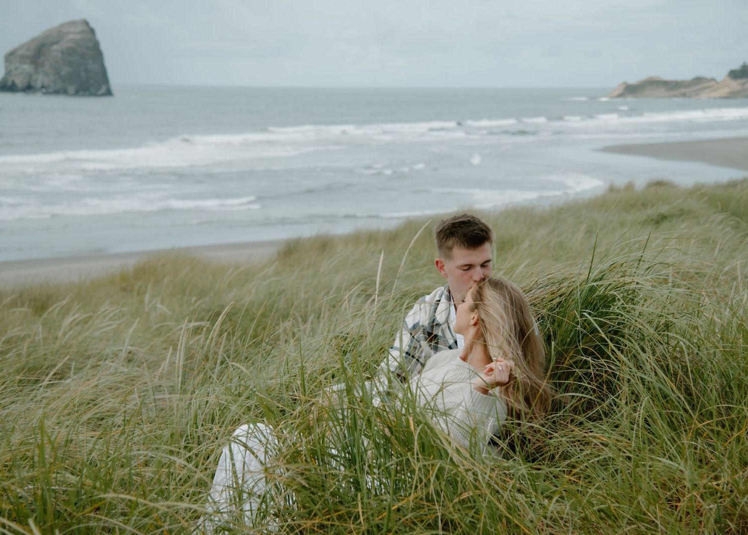 A romantic seaside moment captured in the tall grass dunes with a dramatic coastline in the background on Oregon Coast. Calgary Photographer