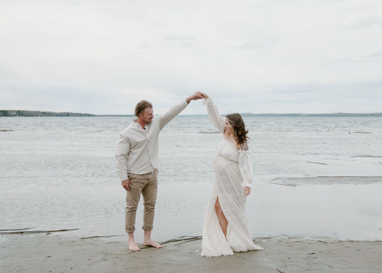 A couple dancing together barefoot on a beach with overcast skies and ocean waves in the background. Calgary Alberta Maternity Photographer.
