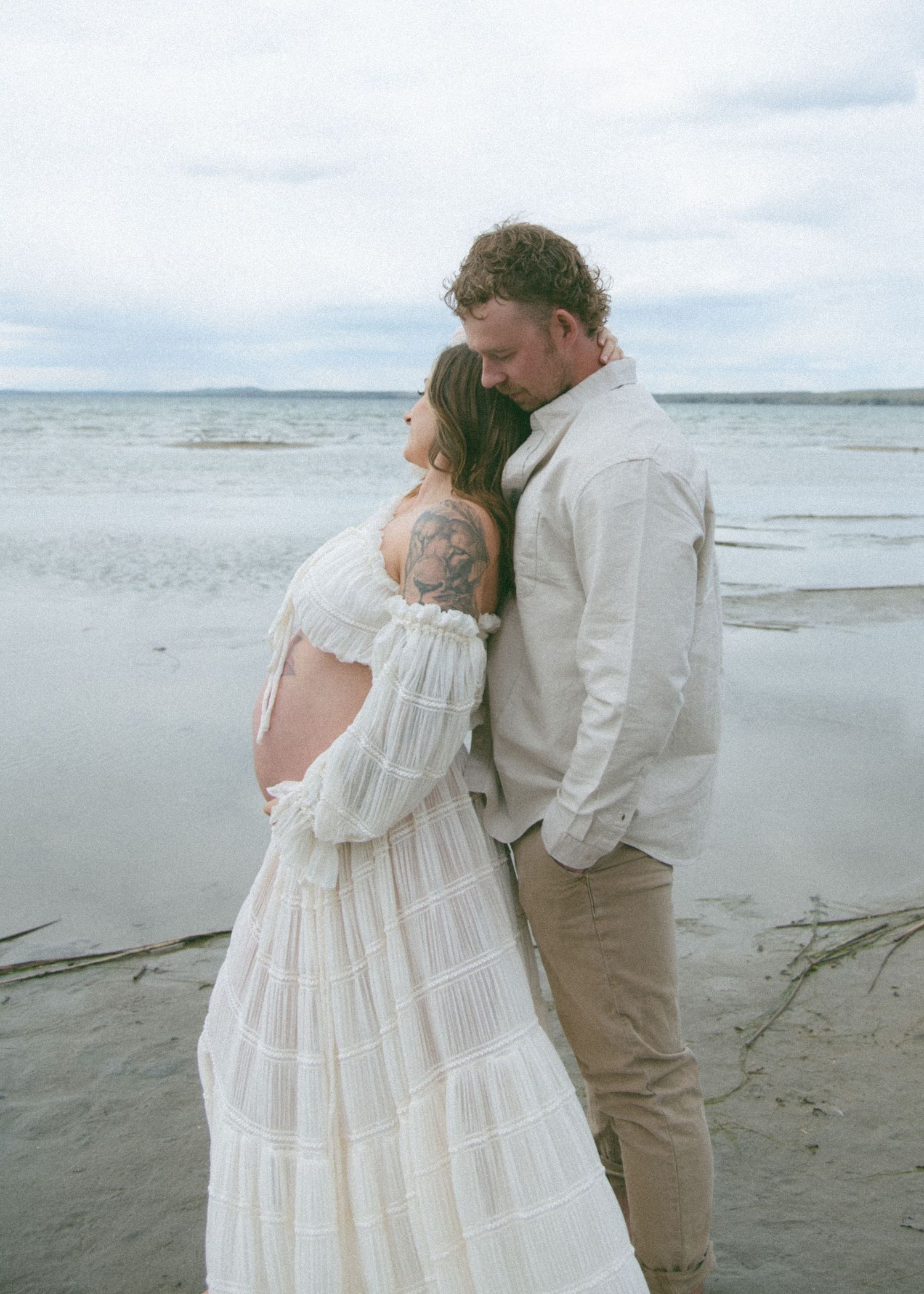 A romantic maternity photoshoot on a cloudy beach showing an intimate moment between a couple in white and beige clothing. Calgary Maternity