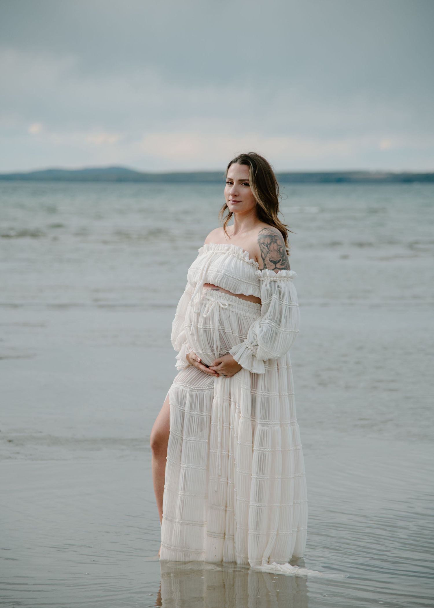 Elegant white gown with delicate details photographed against serene coastal waters. Calgary Alberta Maternity Photographer.