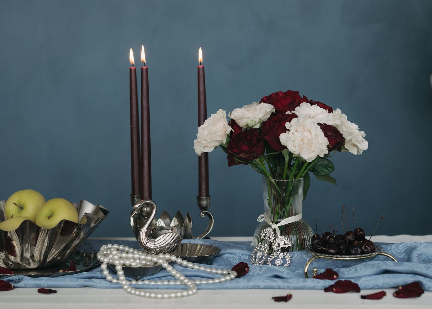 Elegant still life with burgundy and white carnations, silver candlesticks, pearls, and green apples on blue tablecloth.