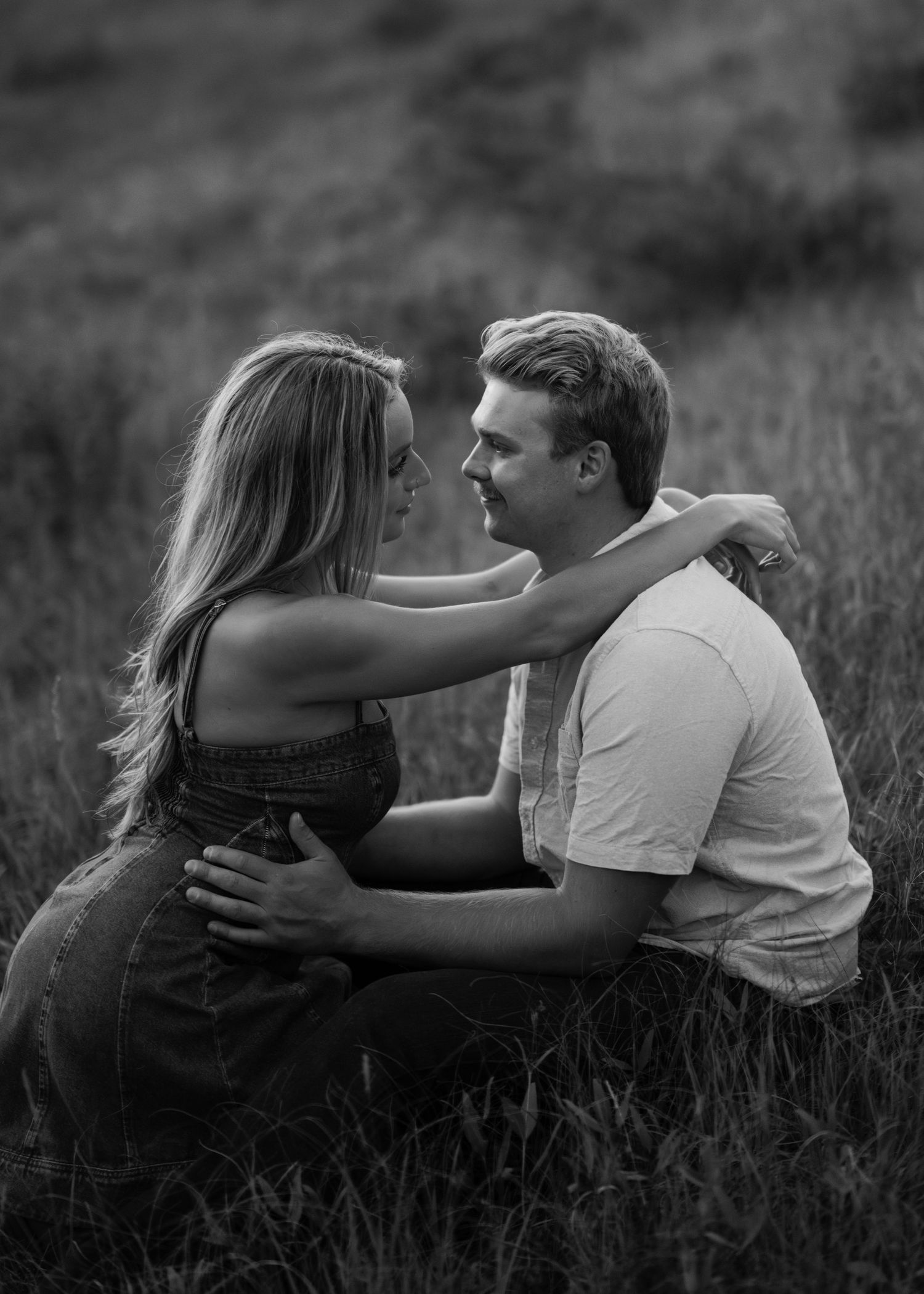 Black and white romantic photoshoot sequence showing a couple embracing and reclining in a grassy meadow at sunset.