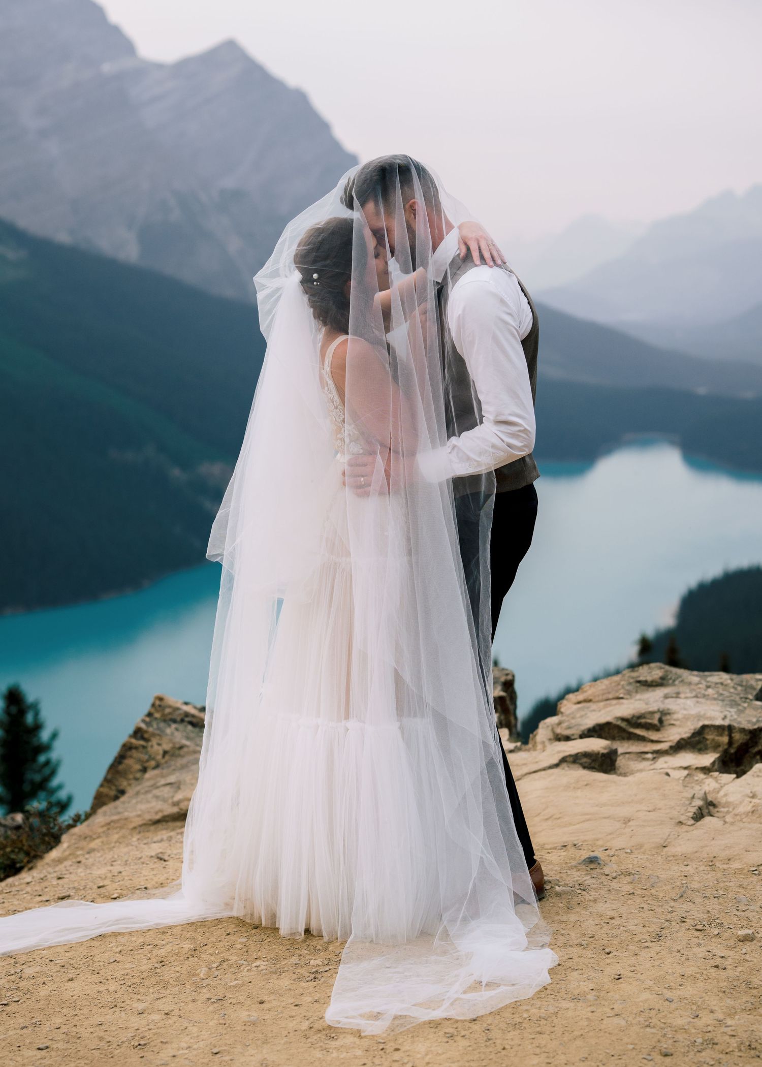 A romantic wedding moment captured on a cliff overlooking a turquoise alpine lake with majestic mountain peaks in the distance.