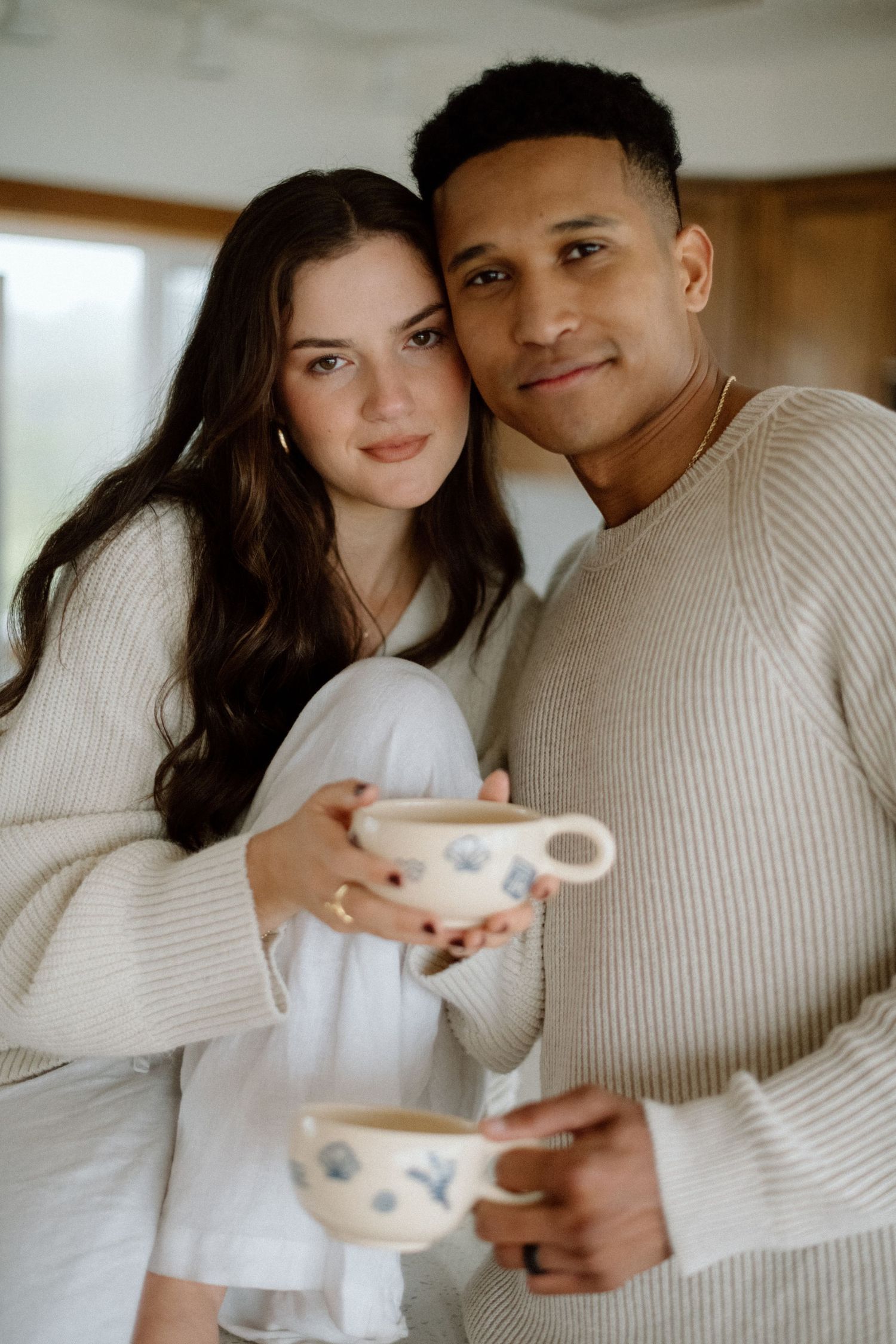 Couple in cozy cream sweaters cuddle on sofa while sharing a warm drink together.