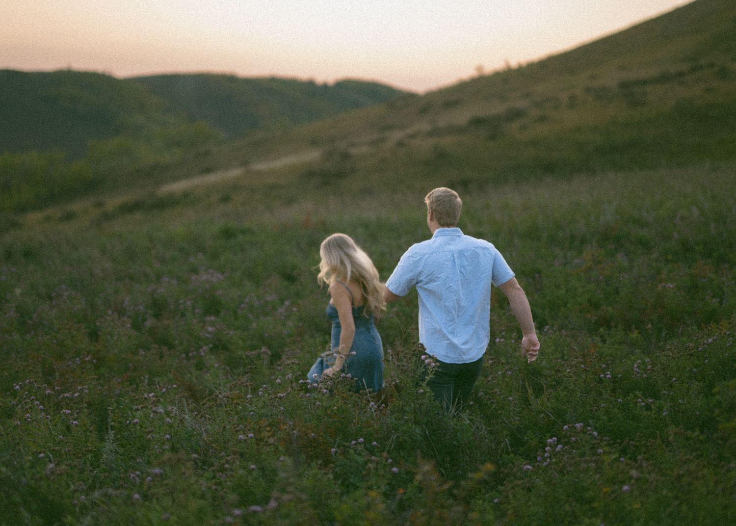 A couple walks together through a scenic hillside meadow at sunset, with long shadows in golden light.