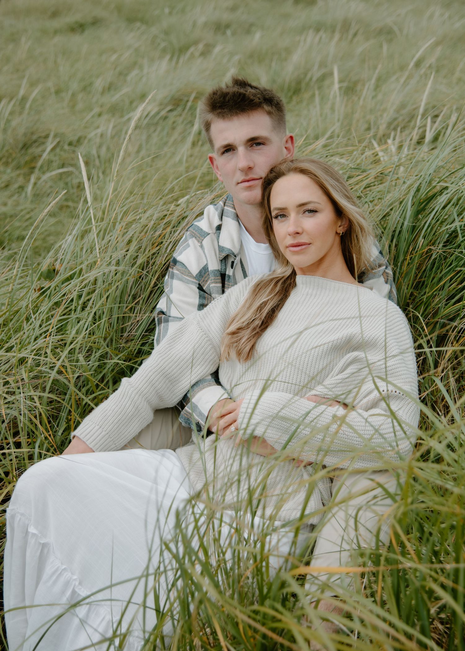 Couple in matching cream sweaters pose together in windswept beach grass on Oregon Coast. Calgary Lifestyle and Engagement Photographer.