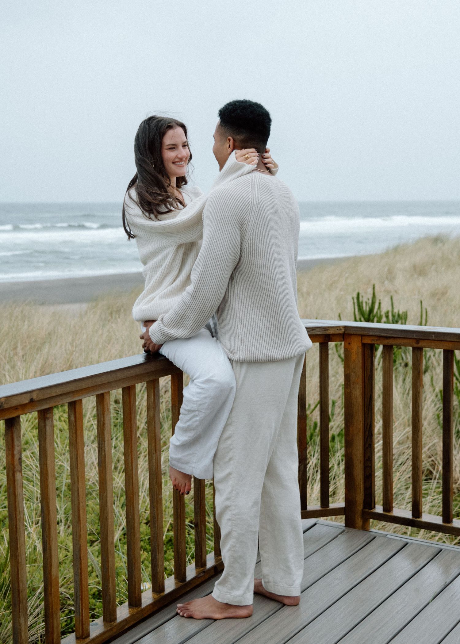 A couple in matching white outfits embraces on a wooden deck overlooking a beach with dune grass and ocean in the background.