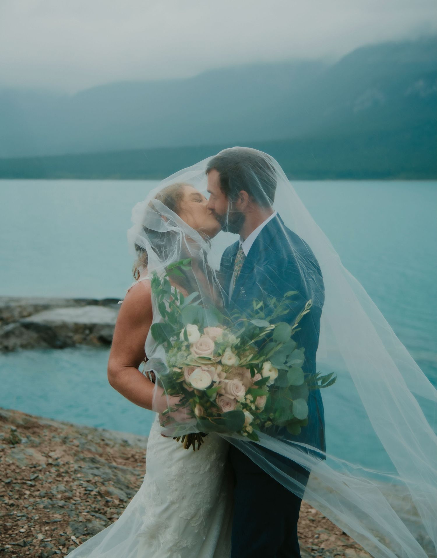 Romantic wedding photos on a rocky lake shore with flowing veil and mountain scenery. Banff and Calgary Wedding Photographer