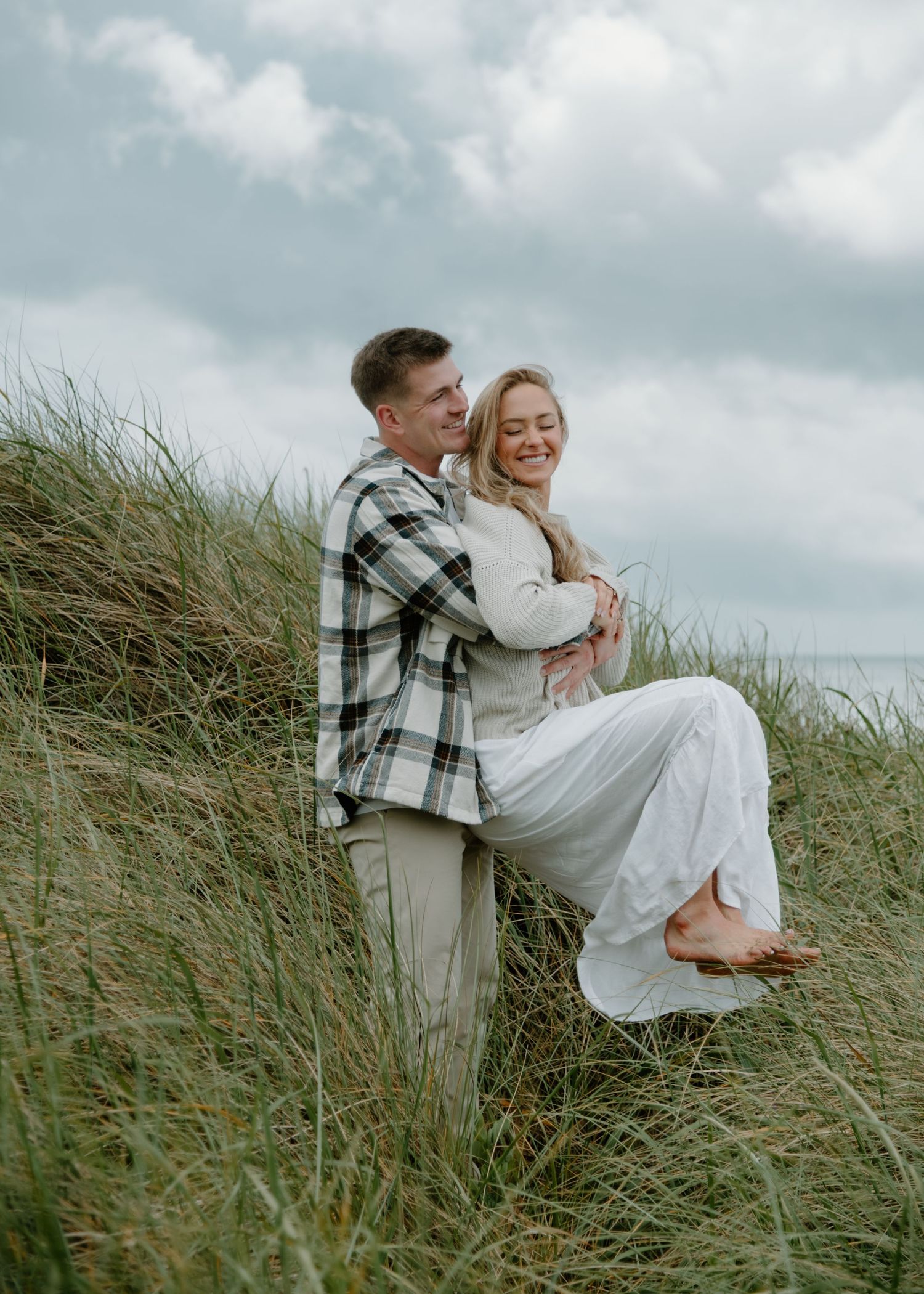 A joyful embrace captured in tall coastal grass against a cloudy sky and ocean backdrop. Calgary Lifestyle and Engagement Photographer