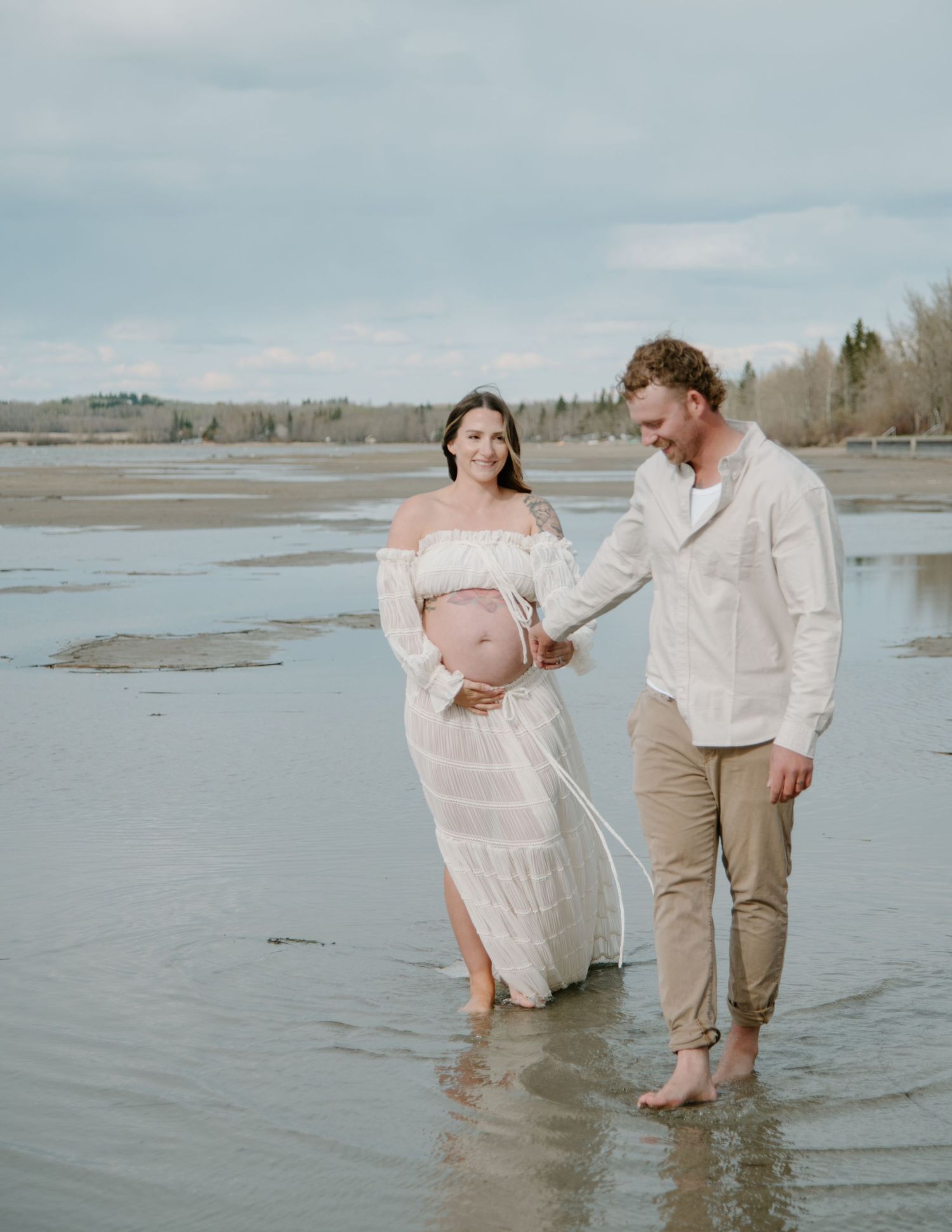 A couple walking barefoot along the shoreline during a beachside maternity photo session. Calgary Alberta Maternity Photographer.