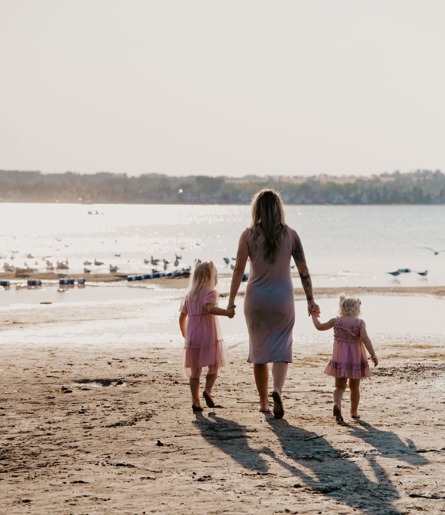 Three figures walk hand-in-hand along a beach shoreline at sunset casting long shadows. Calgary Alberta Family Photographer.