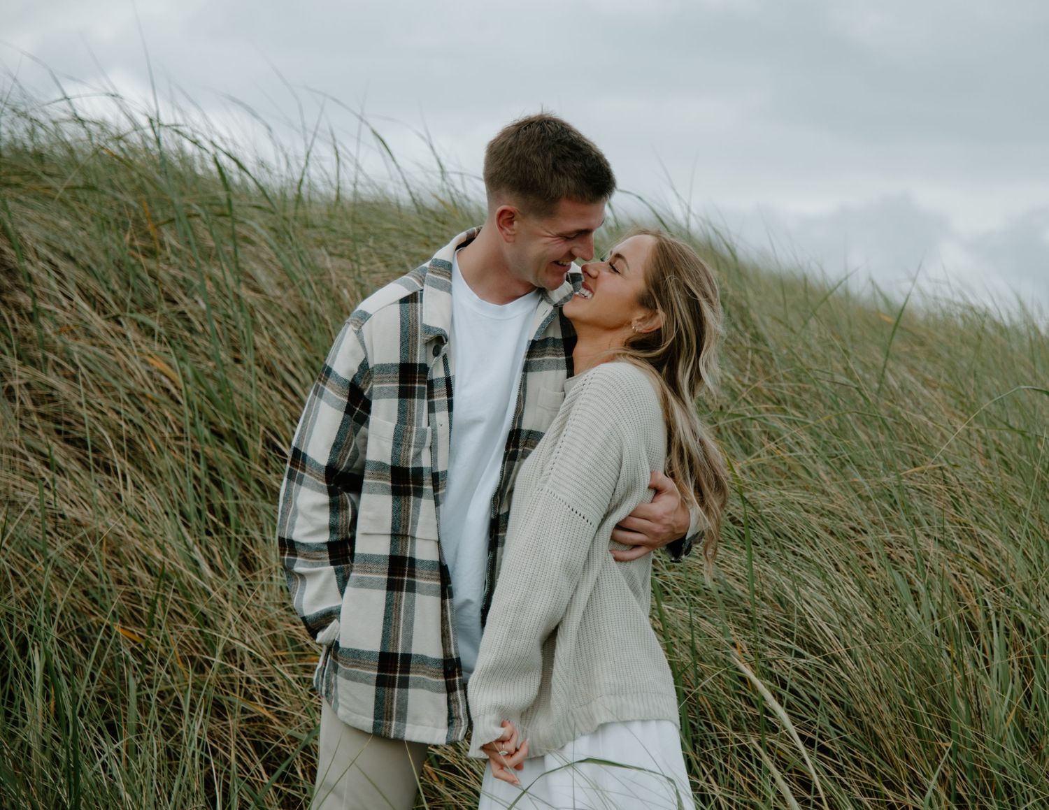 A couple embraces in tall beach grass under overcast skies wearing casual clothing on Oregon Coast. Calgary Lifestyle and Engagement.