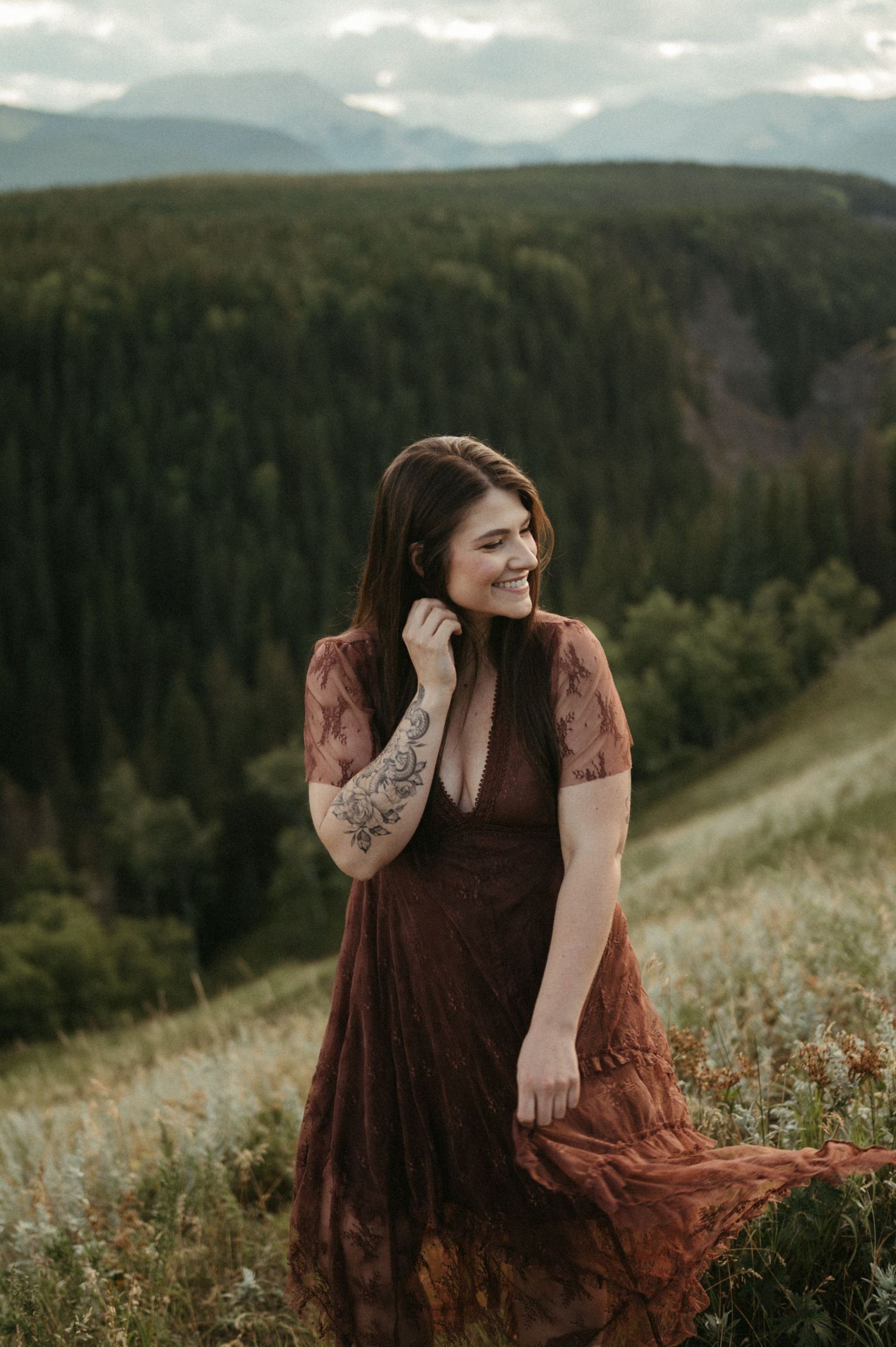 Woman in flowing brown dress standing on mountainside with forest valley view. Calgary Wedding Photographer