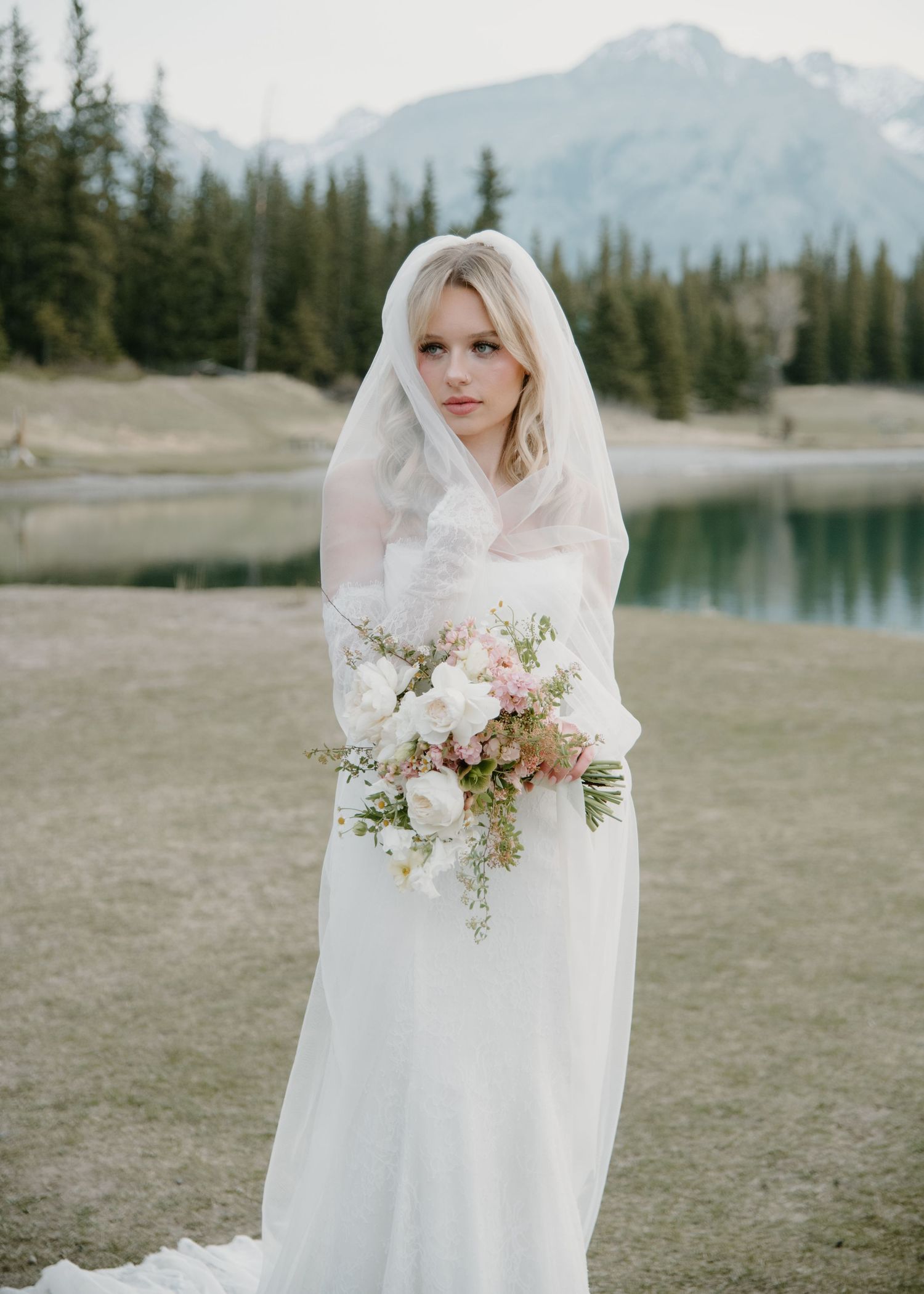 Elegant bridal portrait in simple white dress with delicate bouquet against mountain scenery. Calgary Wedding Photographer