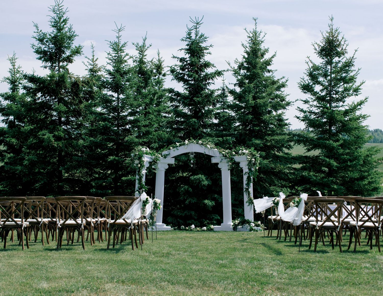 White wooden chairs arranged in rows for an outdoor wedding ceremony with evergreen trees in the background.