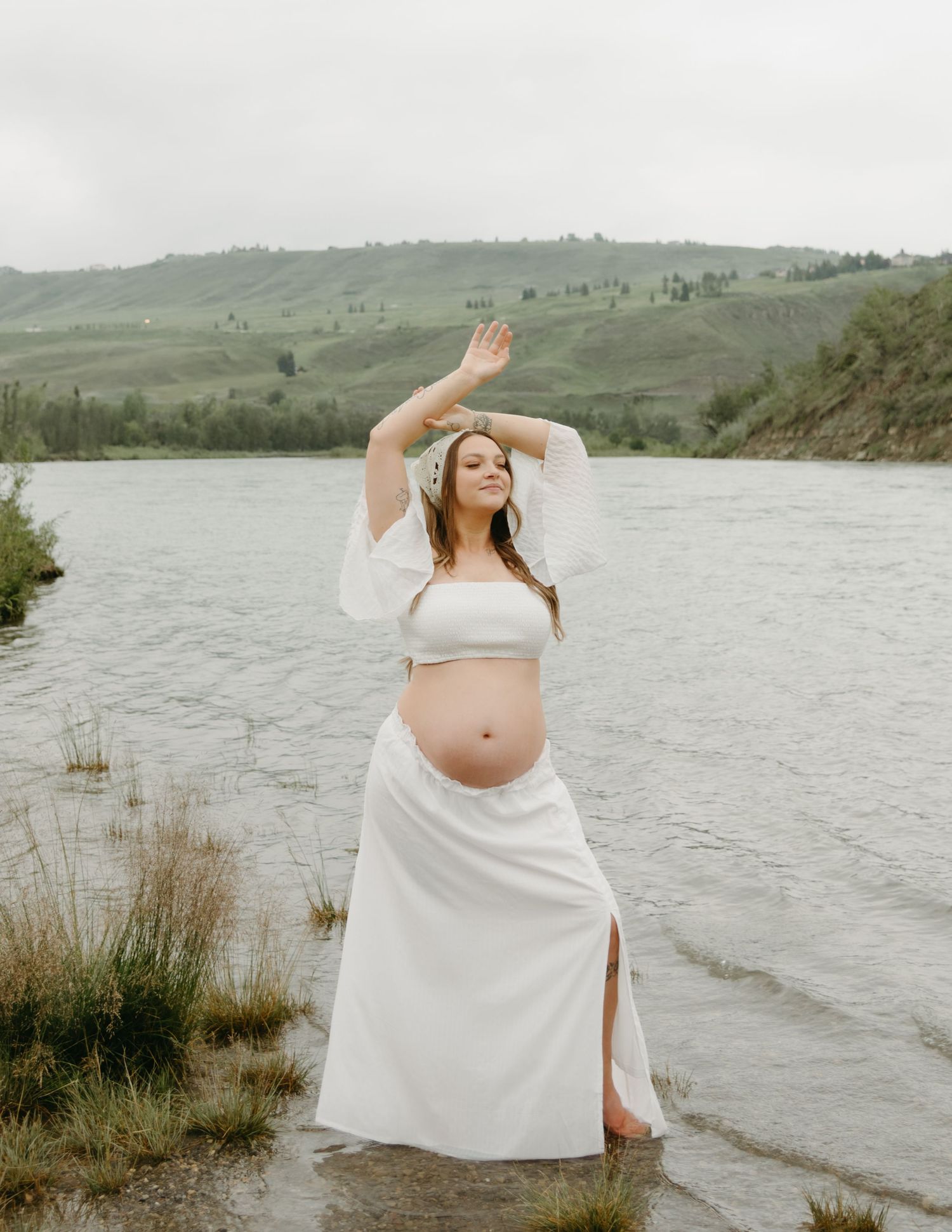 Ethereal maternity photoshoot by a lake with rolling green hills in the background featuring flowing white fabric.