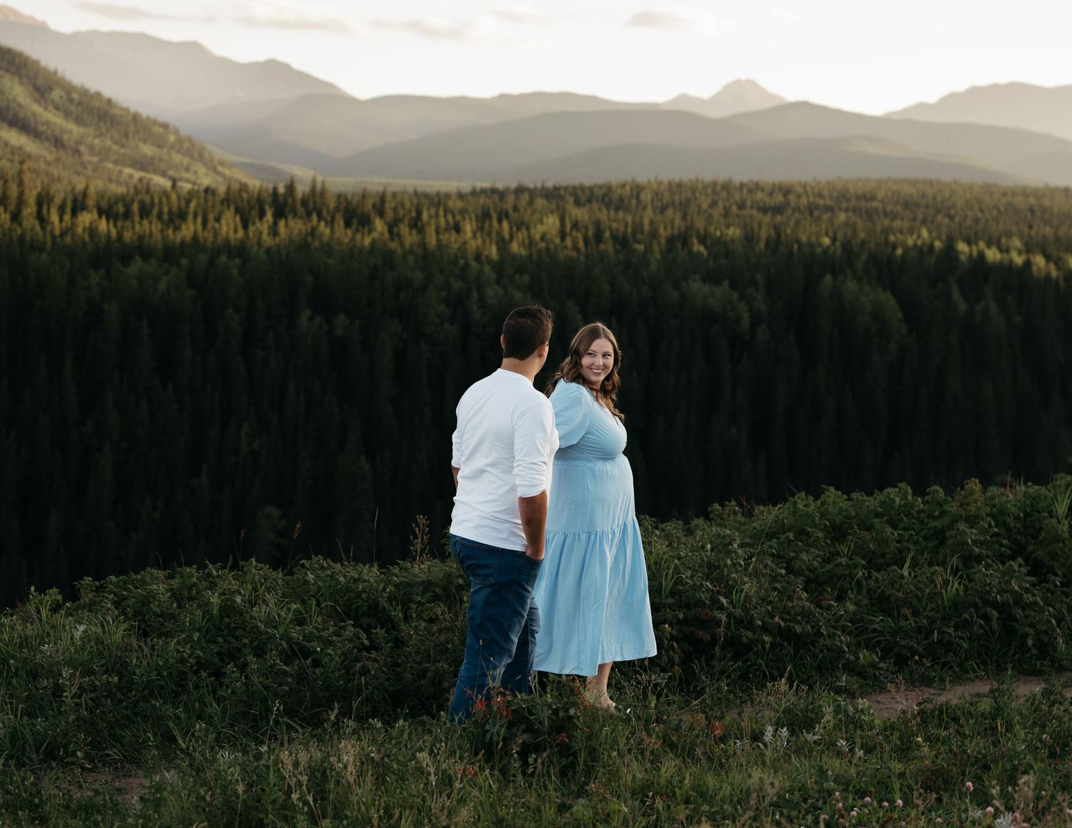 Two people standing on a mountainside together overlooking a scenic forest landscape during golden hour.