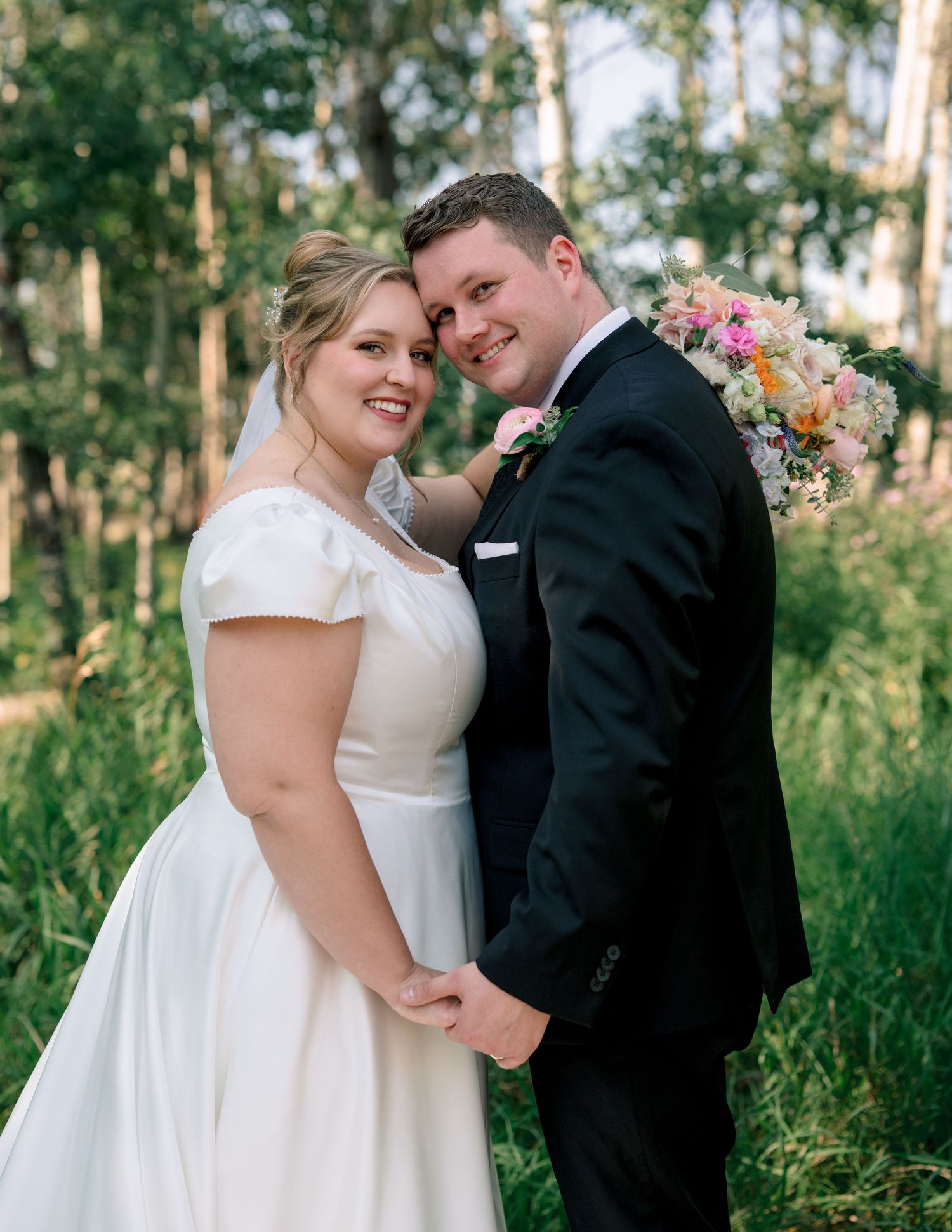 A newly married couple embraces in a sun-dappled forest clearing with a colorful floral bouquet.