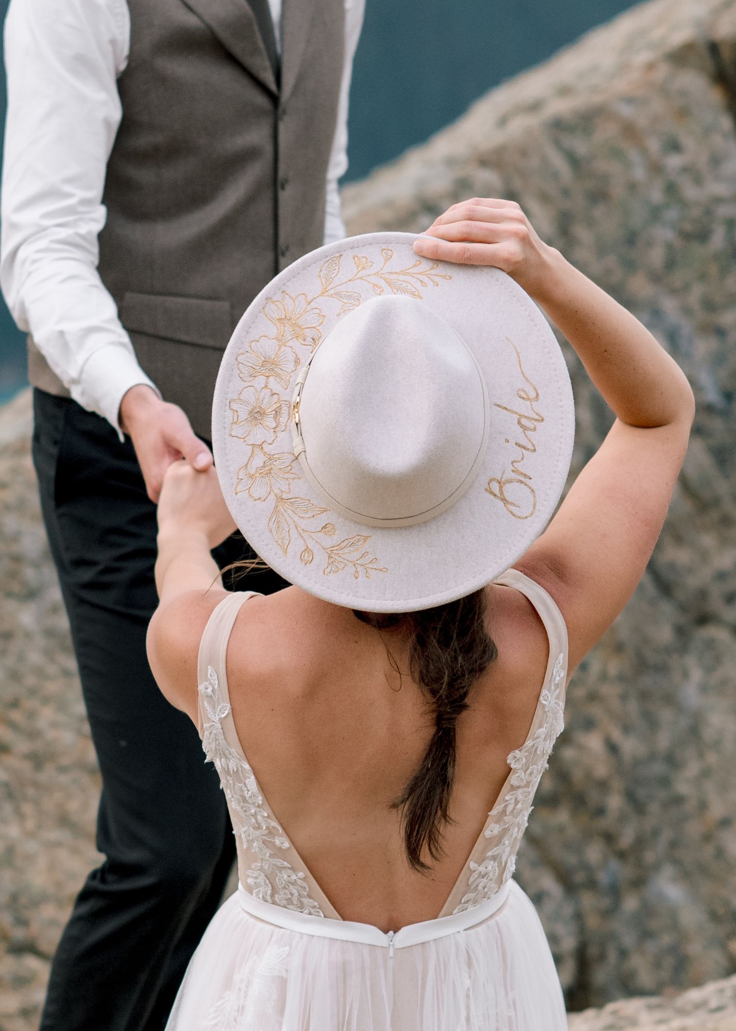 A bride in a white wedding dress and sun hat rests on coastal rocks during an outdoor beach photo session.