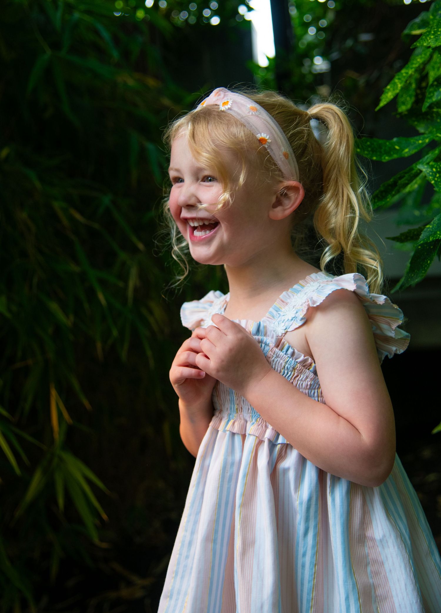 Child in blue and white striped dress laughs with pure joy among green foliage. Calgary Alberta Family Photographer.