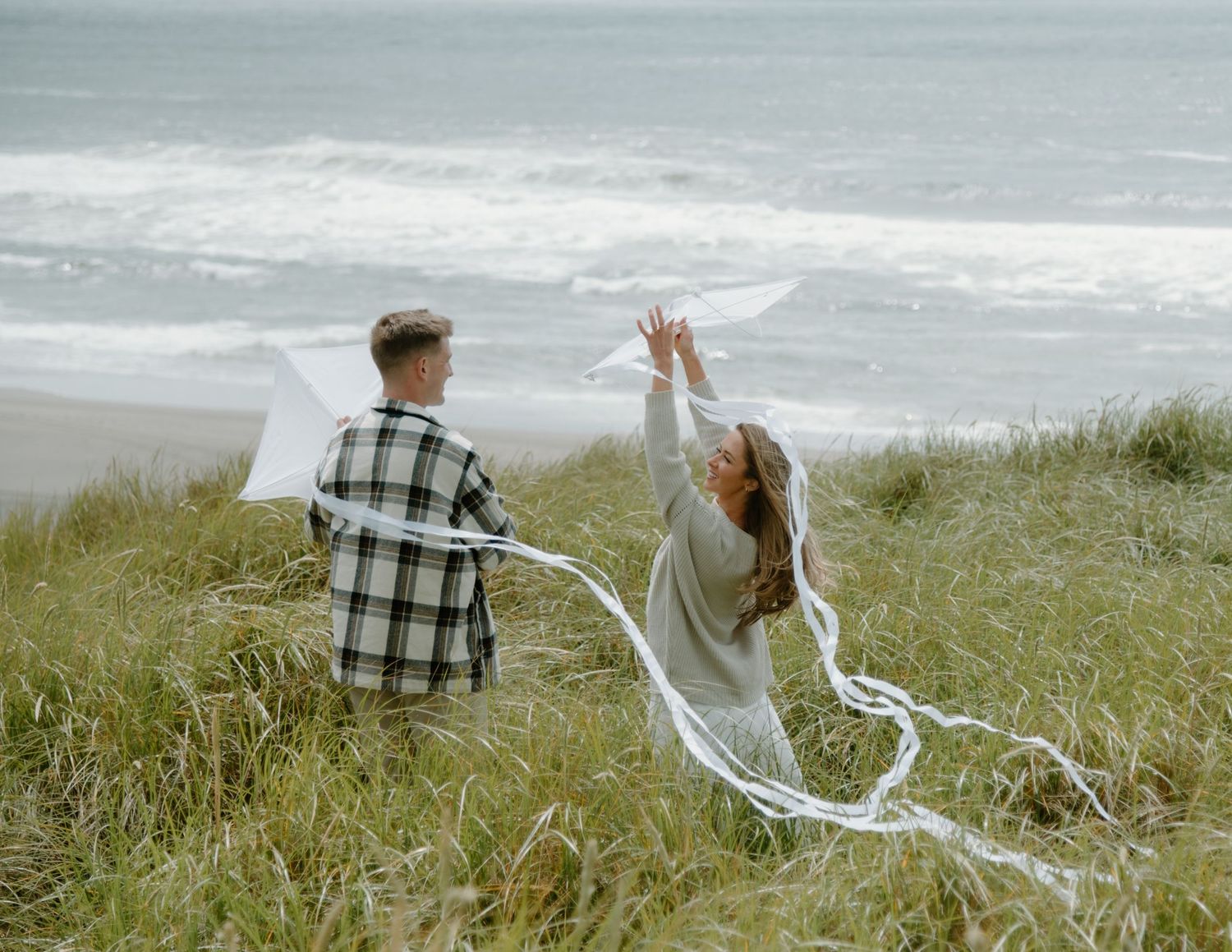 A couple celebrates on a grassy coastal hillside with flowing white fabric on Oregon Coast. Calgary Lifestyle and Engagement Photographer.