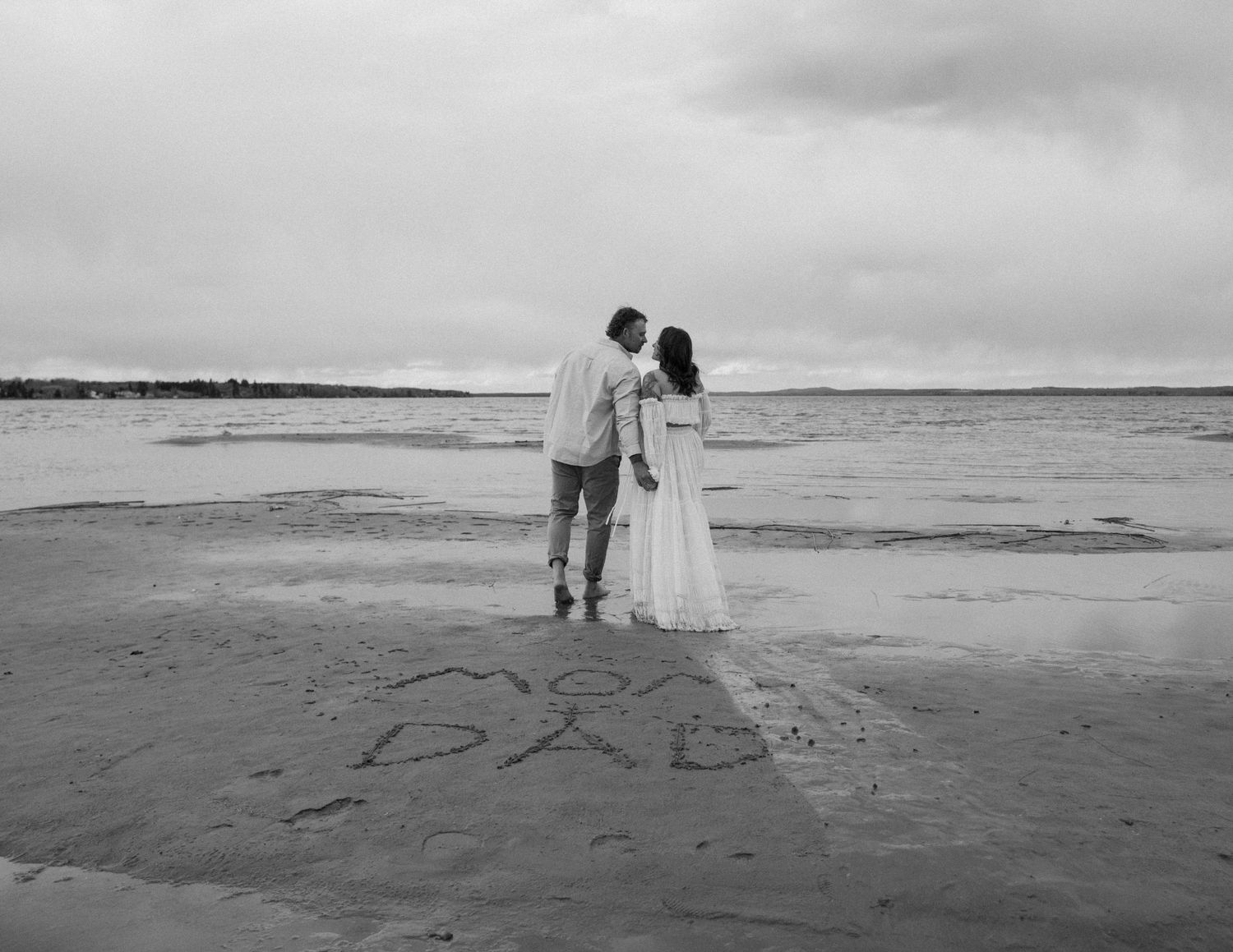 A black and white photo of a couple standing on beach with writing in sand. Calgary Alberta Maternity Photographer.