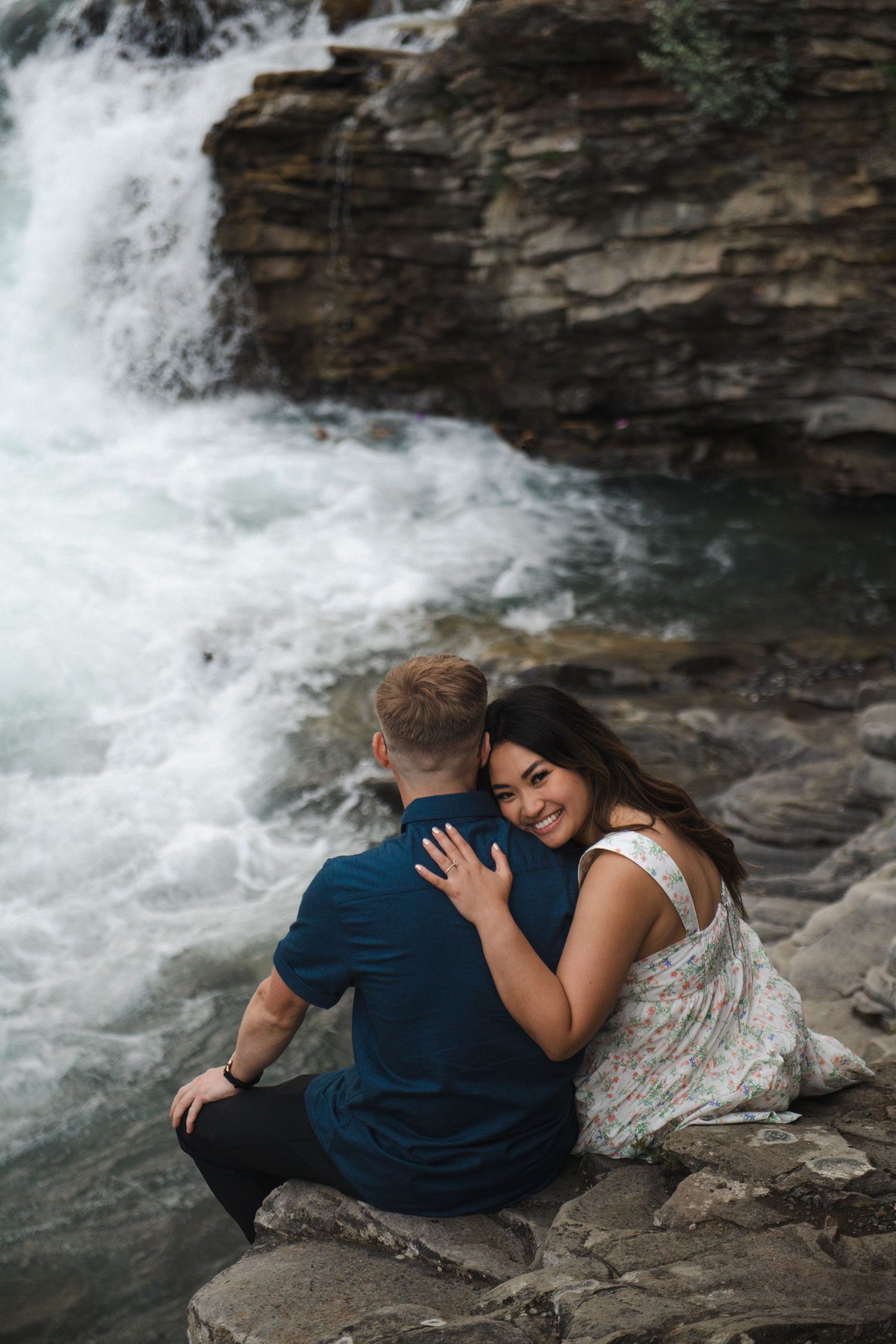 Couple resting together on rocky ledge beside rushing waterfall in Calgary. Calgary Lifestyle and Engagement Photographer.