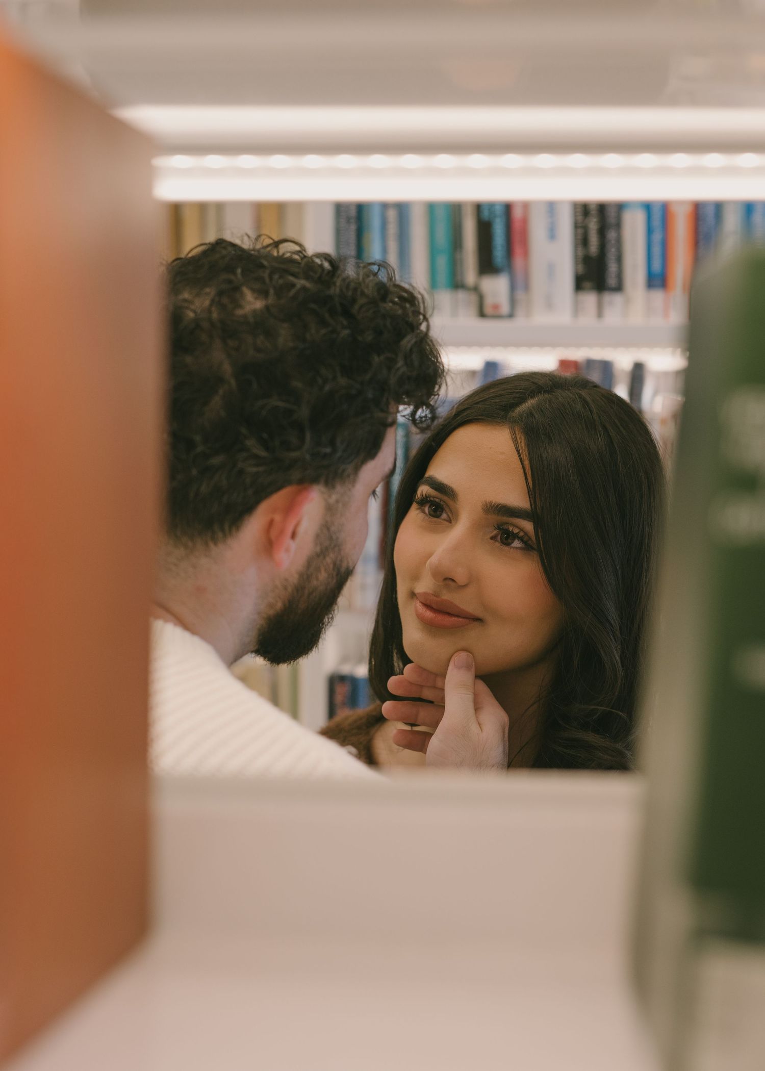 A romantic couple shares an intimate moment in a library setting, framed through bookshelves and soft lighting.