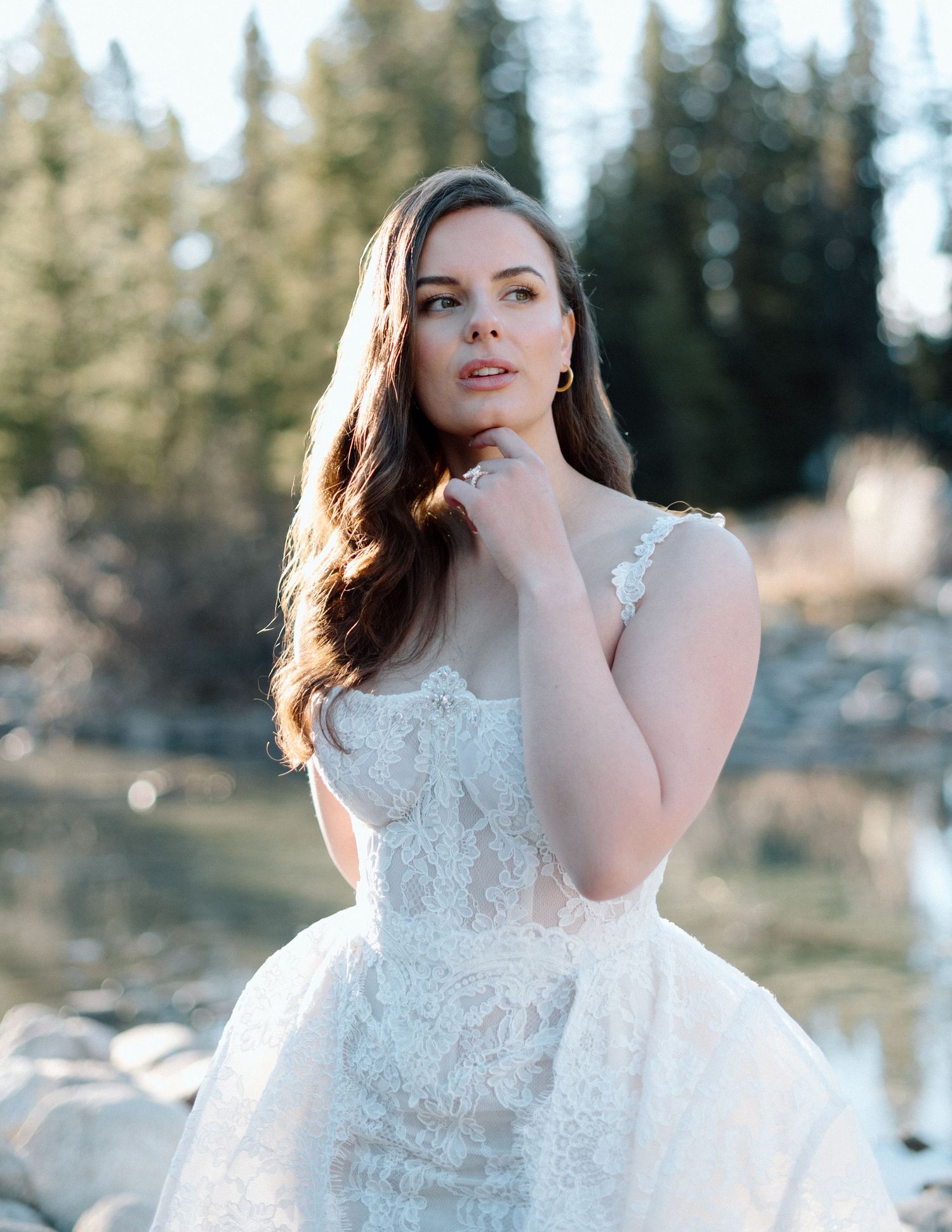 A bride in a delicate lace wedding gown poses outdoors against a backdrop of evergreen trees at Lake Louise. Calgary Wedding Photographer