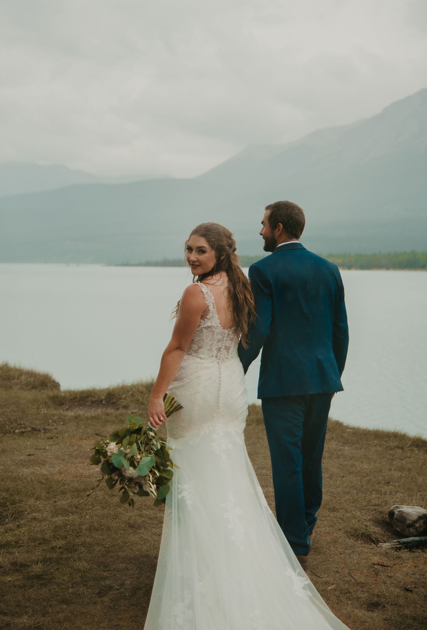 Elegant wedding couple poses on cliff edge overlooking serene mountain lake. Calgary Wedding Photographer