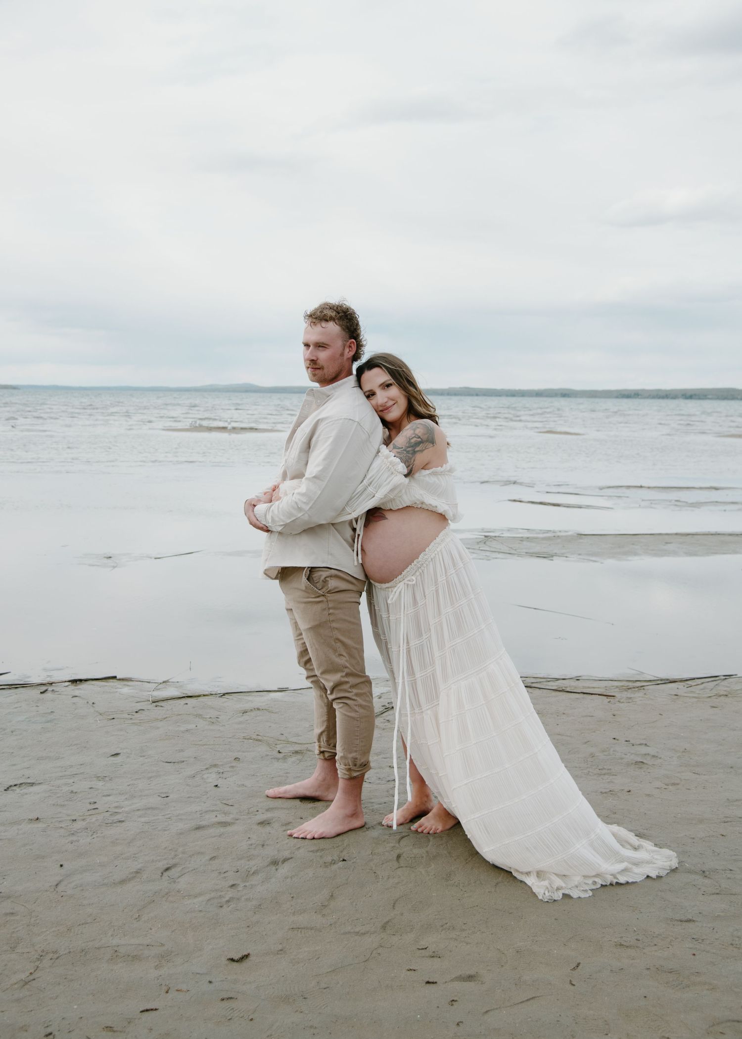 A couple embraces during a romantic beach photoshoot with waves and cloudy skies in the background. Calgary Alberta Maternity Photographer.