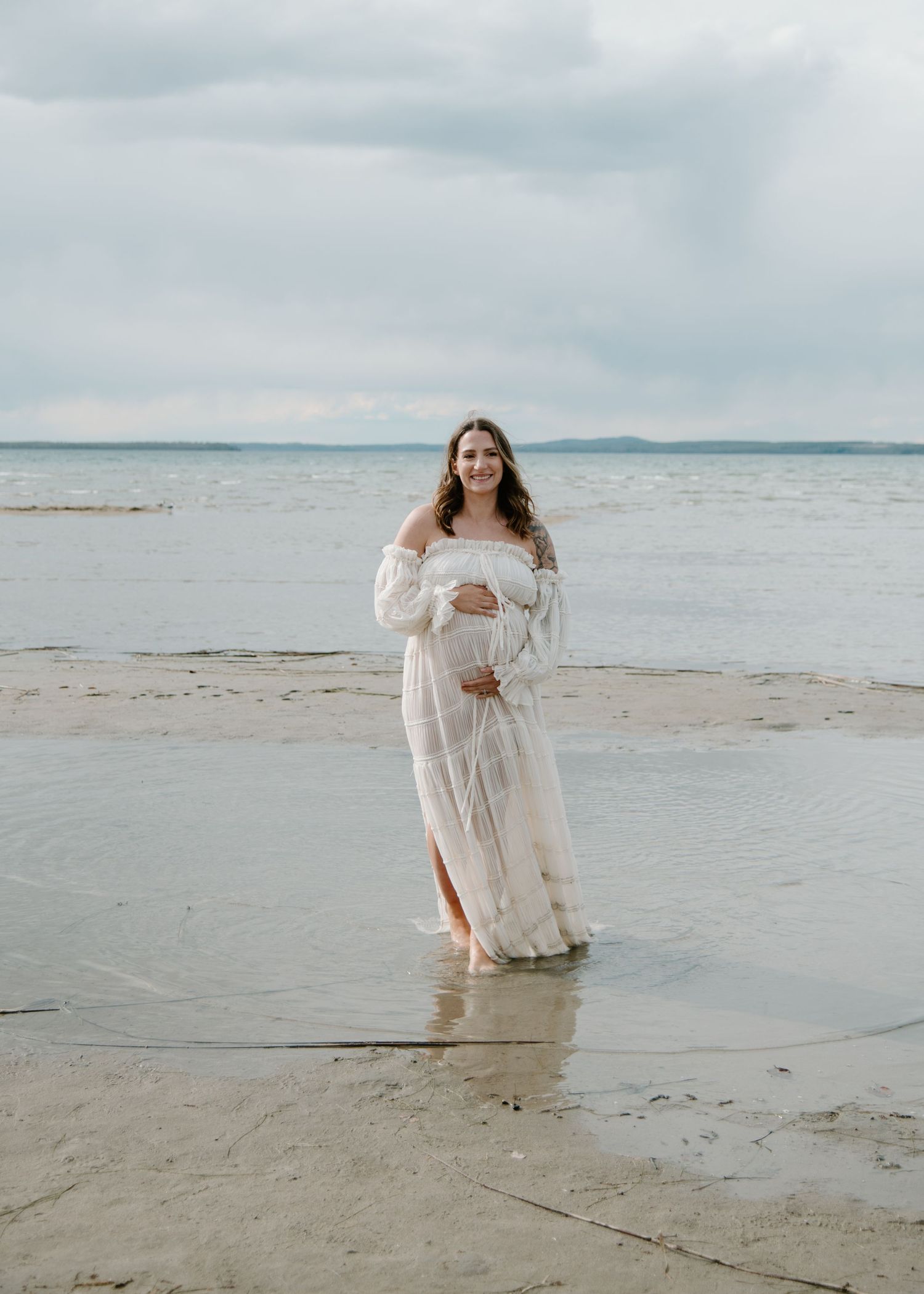 Beachwear maternity portrait with flowing white dress and cloudy coastal background. Calgary Alberta Maternity Photographer.