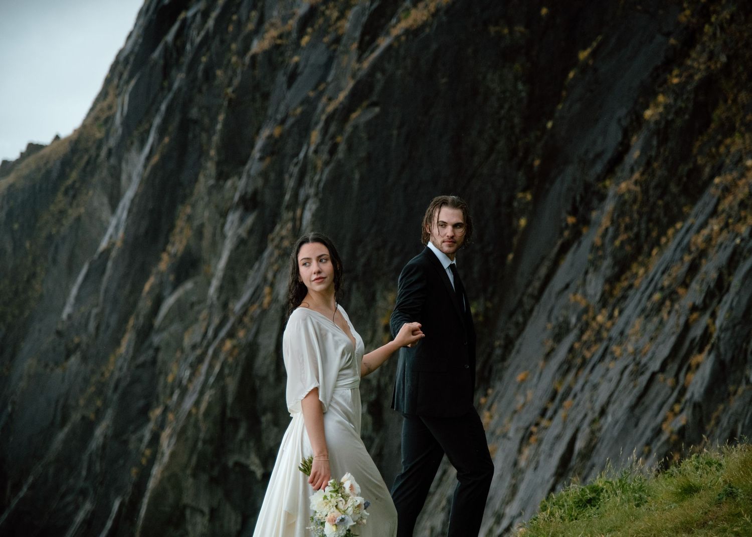 A wedding couple poses elegantly against towering coastal cliffs with dramatic natural lighting. Calgary Wedding Photographer