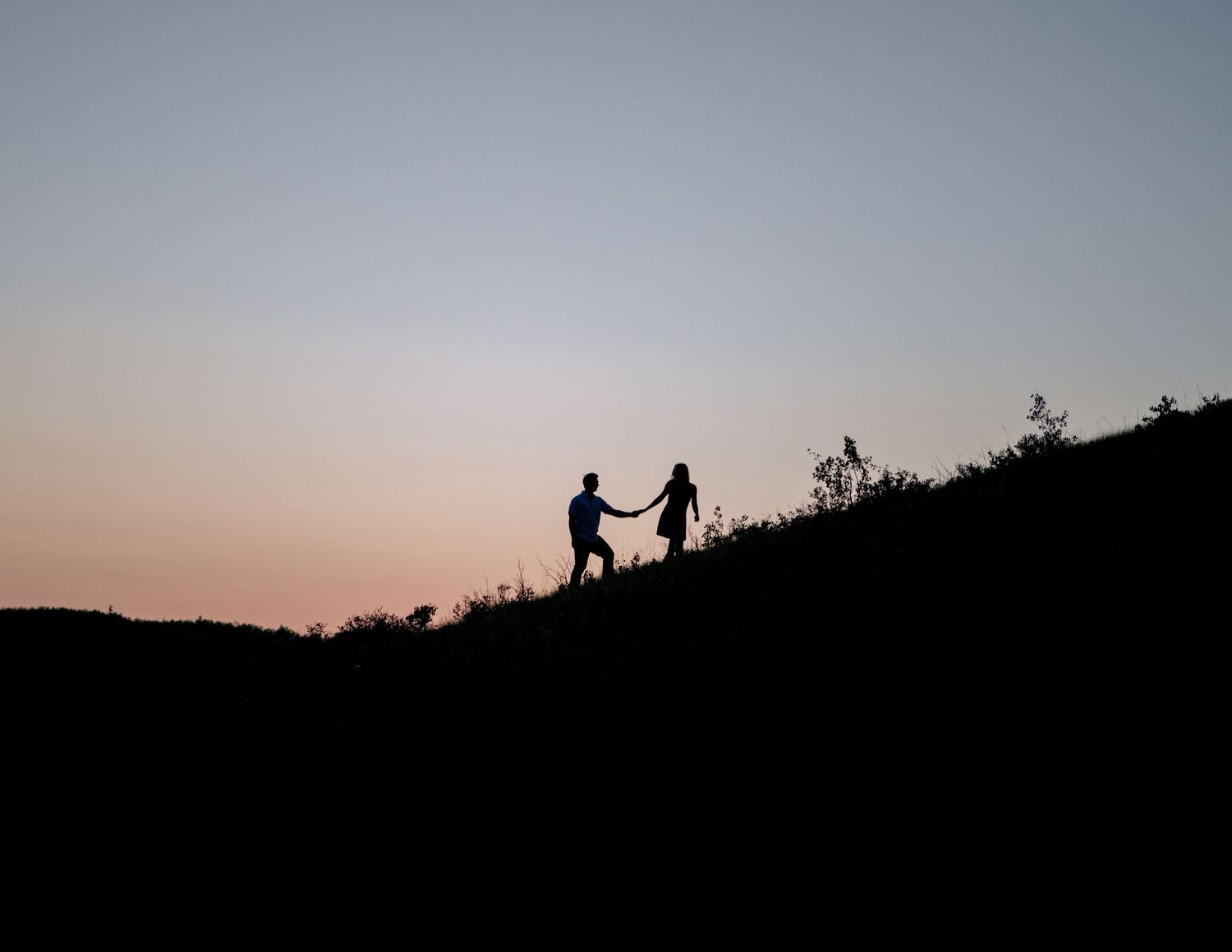 Two figures hold hands in silhouette against a sunset sky as they walk along a mountain ridge.